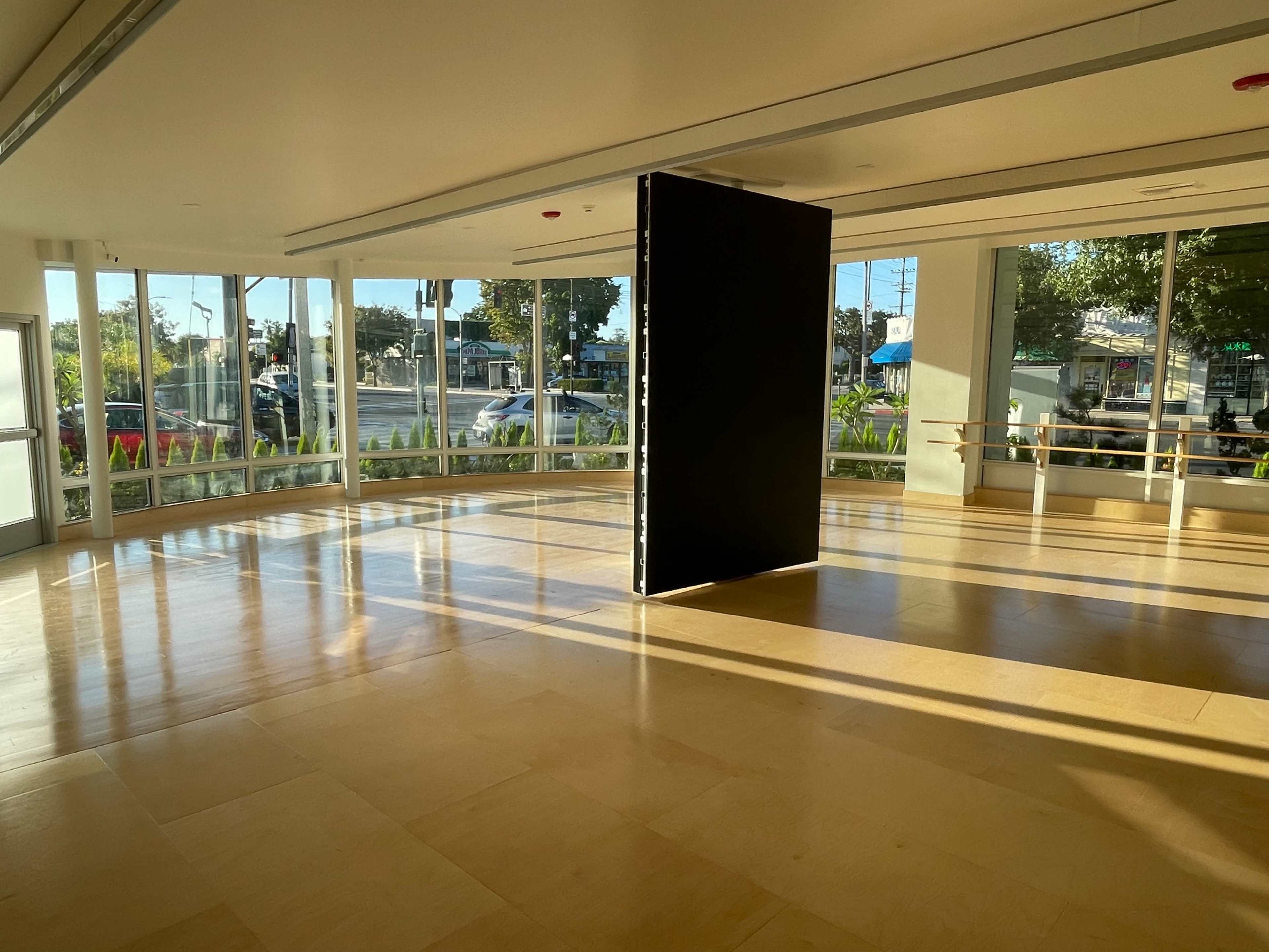 The image shows an empty room with large windows, a black partition in the center, and sunlight casting shadows on the floor.