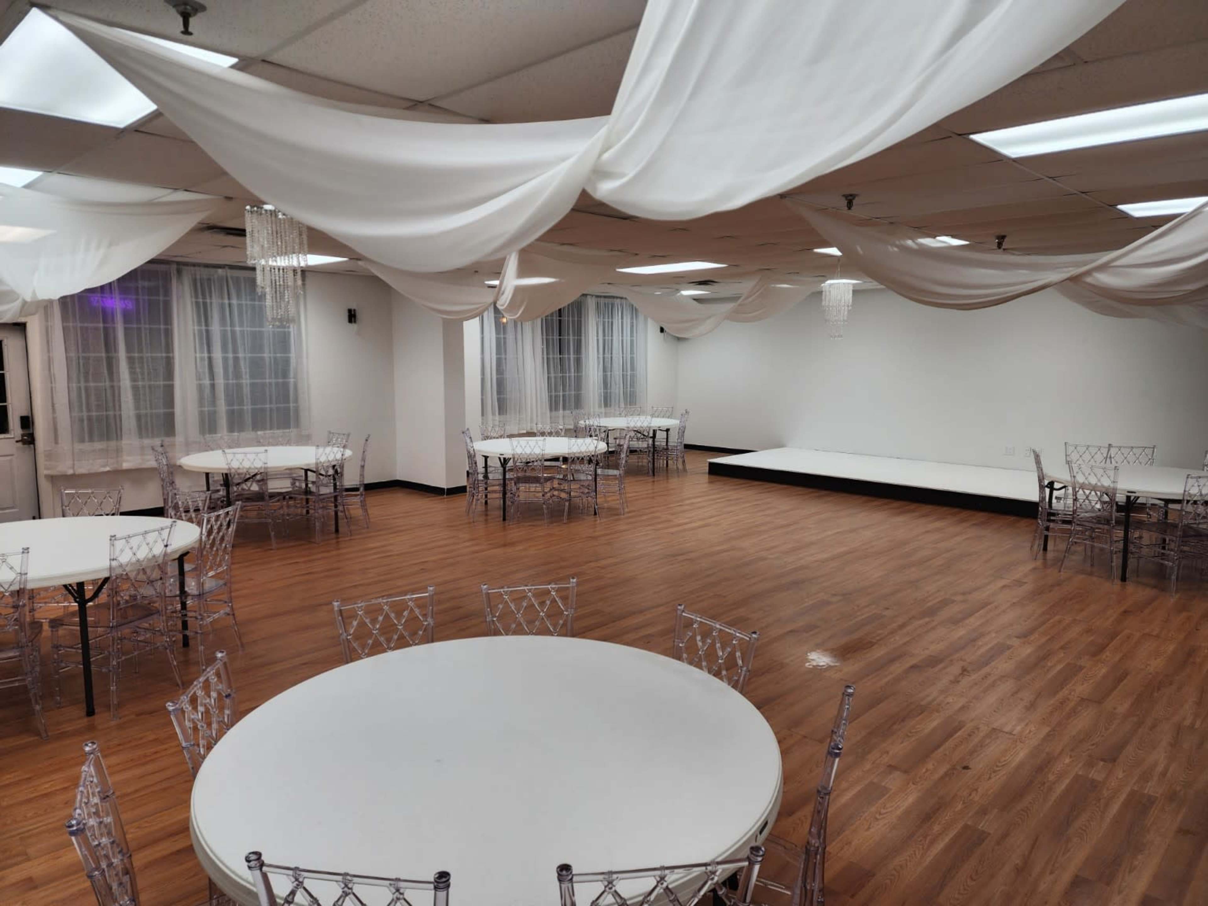 The room features a spacious layout with wooden flooring, circular tables surrounded by clear acrylic chairs, and draped white fabric hanging from the ceiling.