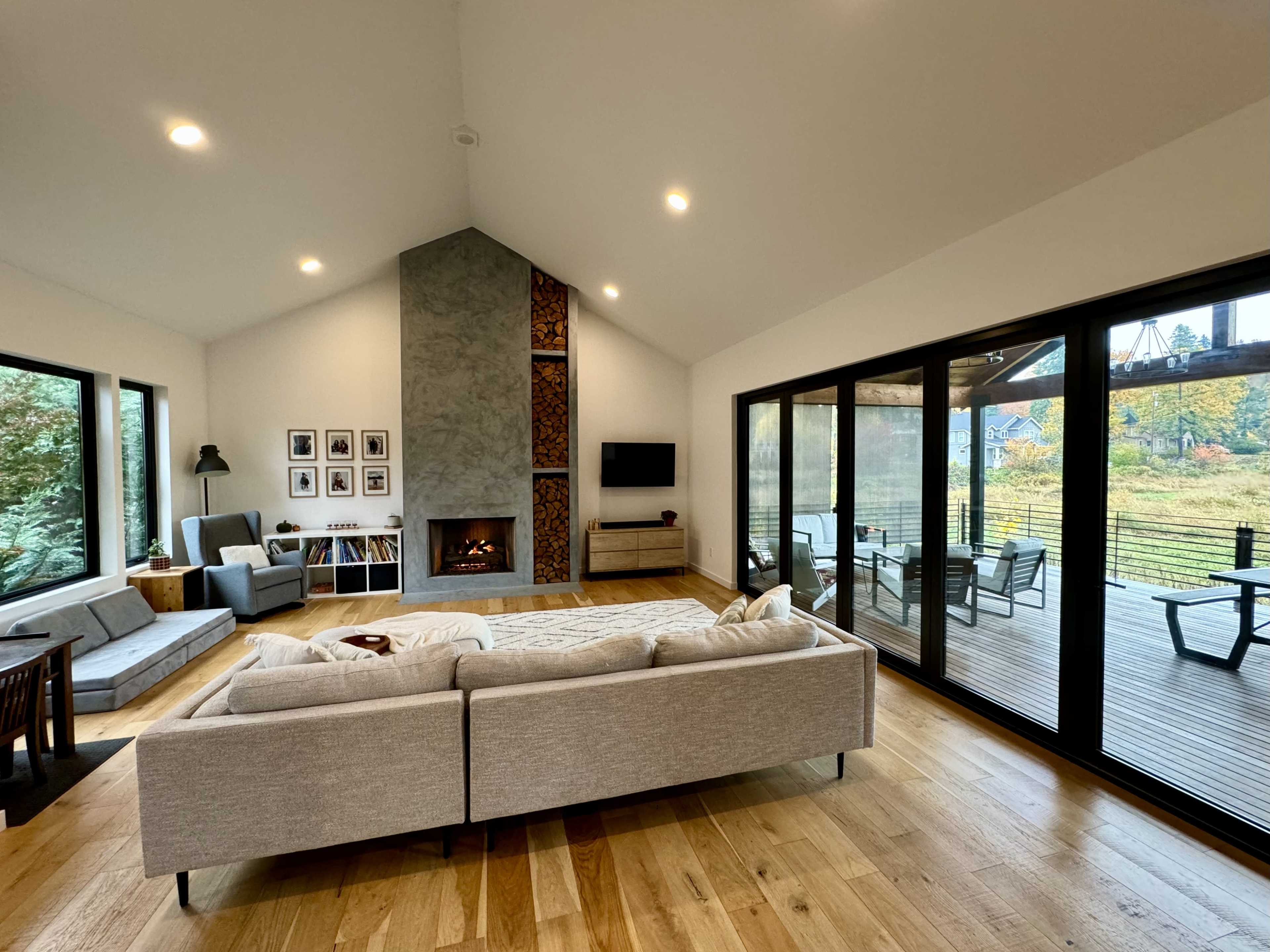A modern living room features a large gray sofa, a fireplace with a stone facade, and expansive glass doors that open to a wooden deck.