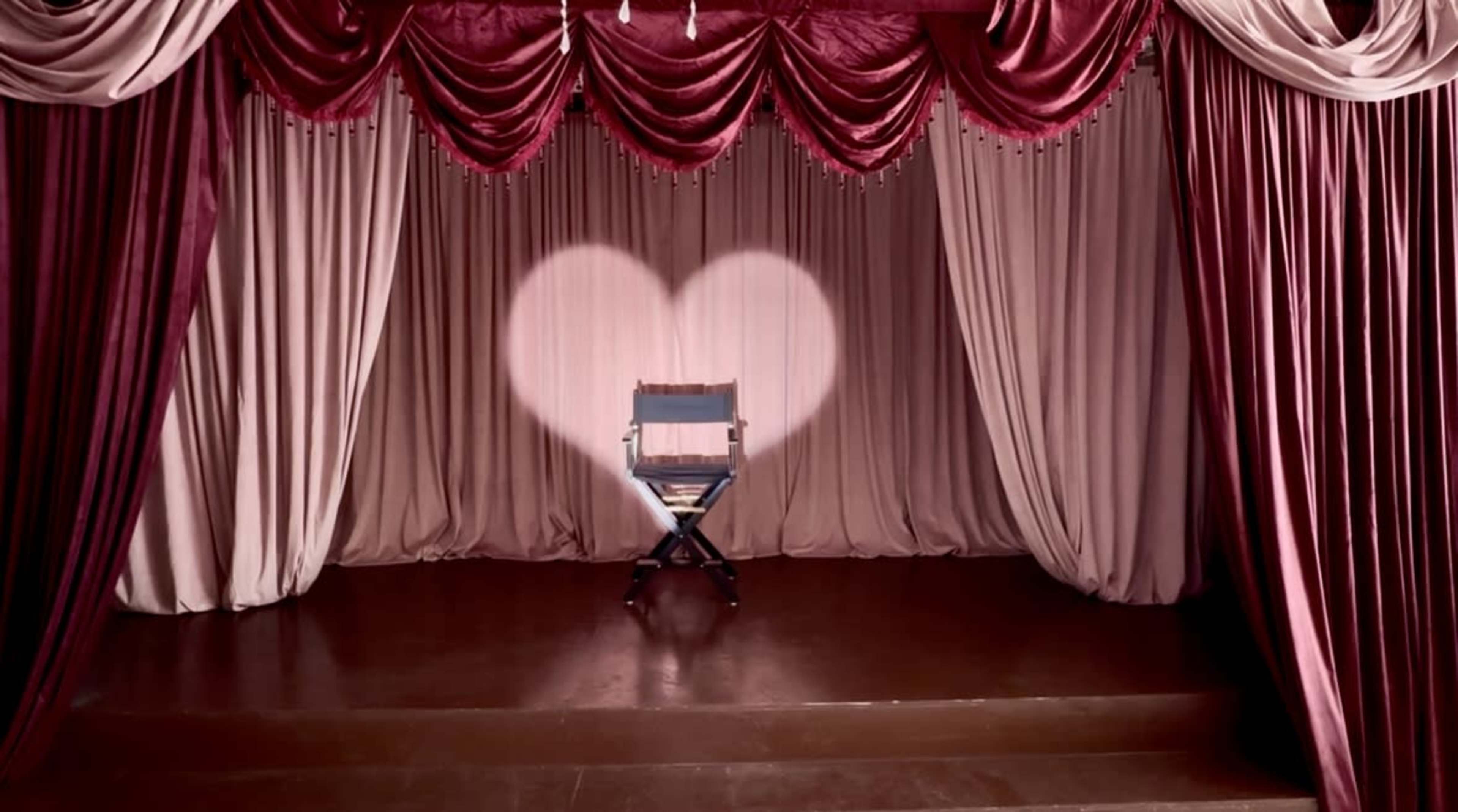 A spotlight forms a heart shape on the floor in front of an empty director's chair, surrounded by draped curtains in shades of pink and red.