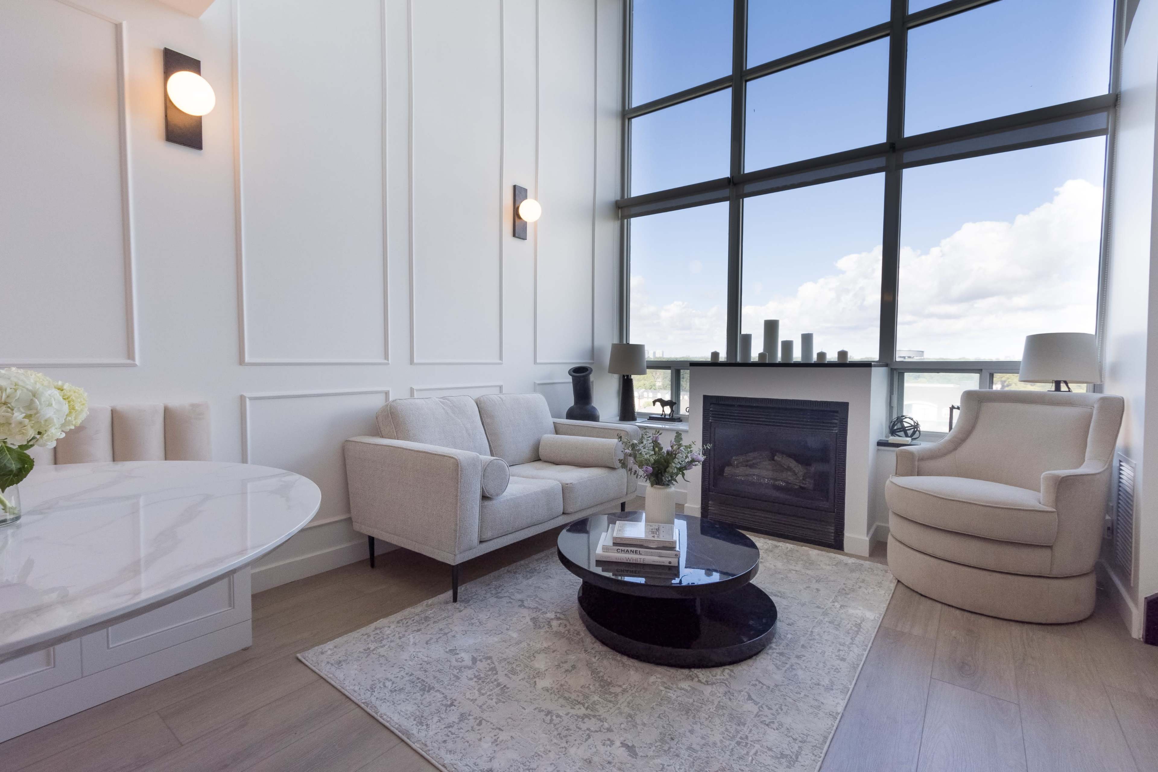 A modern living room features a light-colored sofa, a round coffee table, an armchair, and a fireplace beneath large windows showcasing a view of the sky.