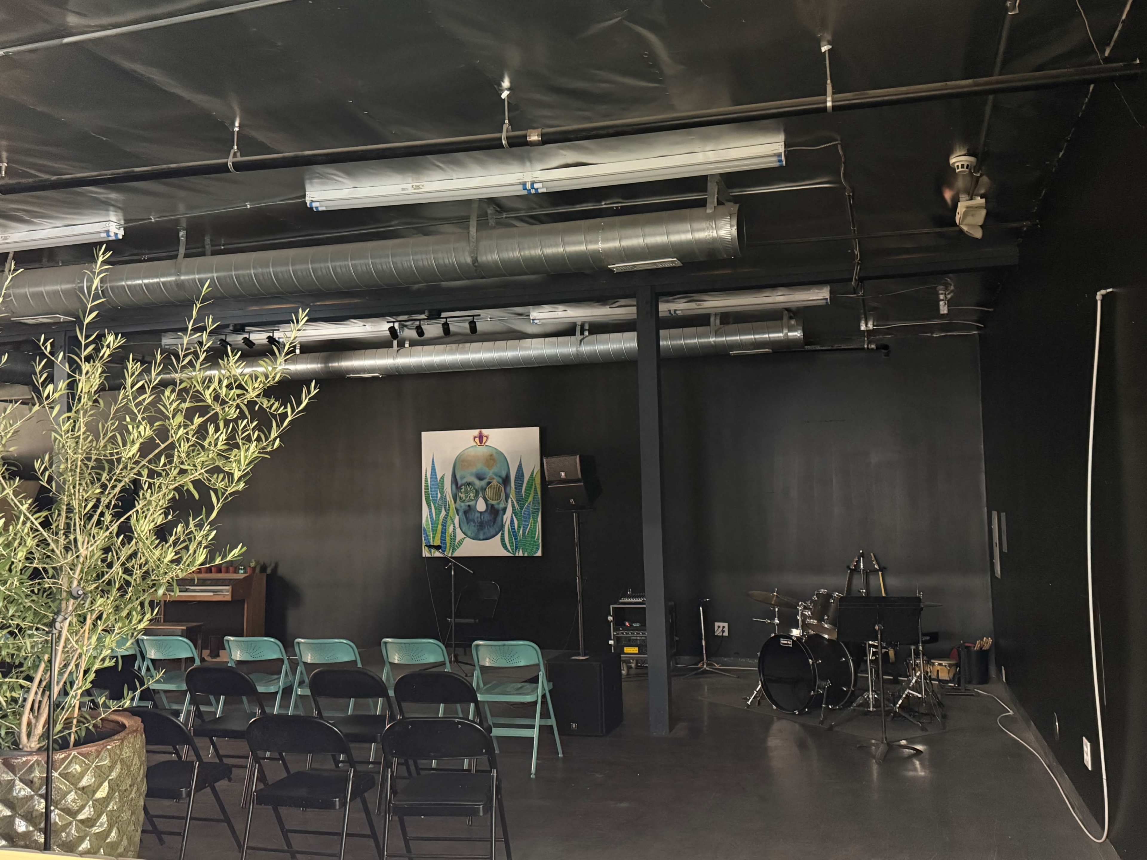 The image shows a dimly lit room featuring a small stage with a drum set, chairs arranged for an audience, and a large artwork of a skull on the wall.