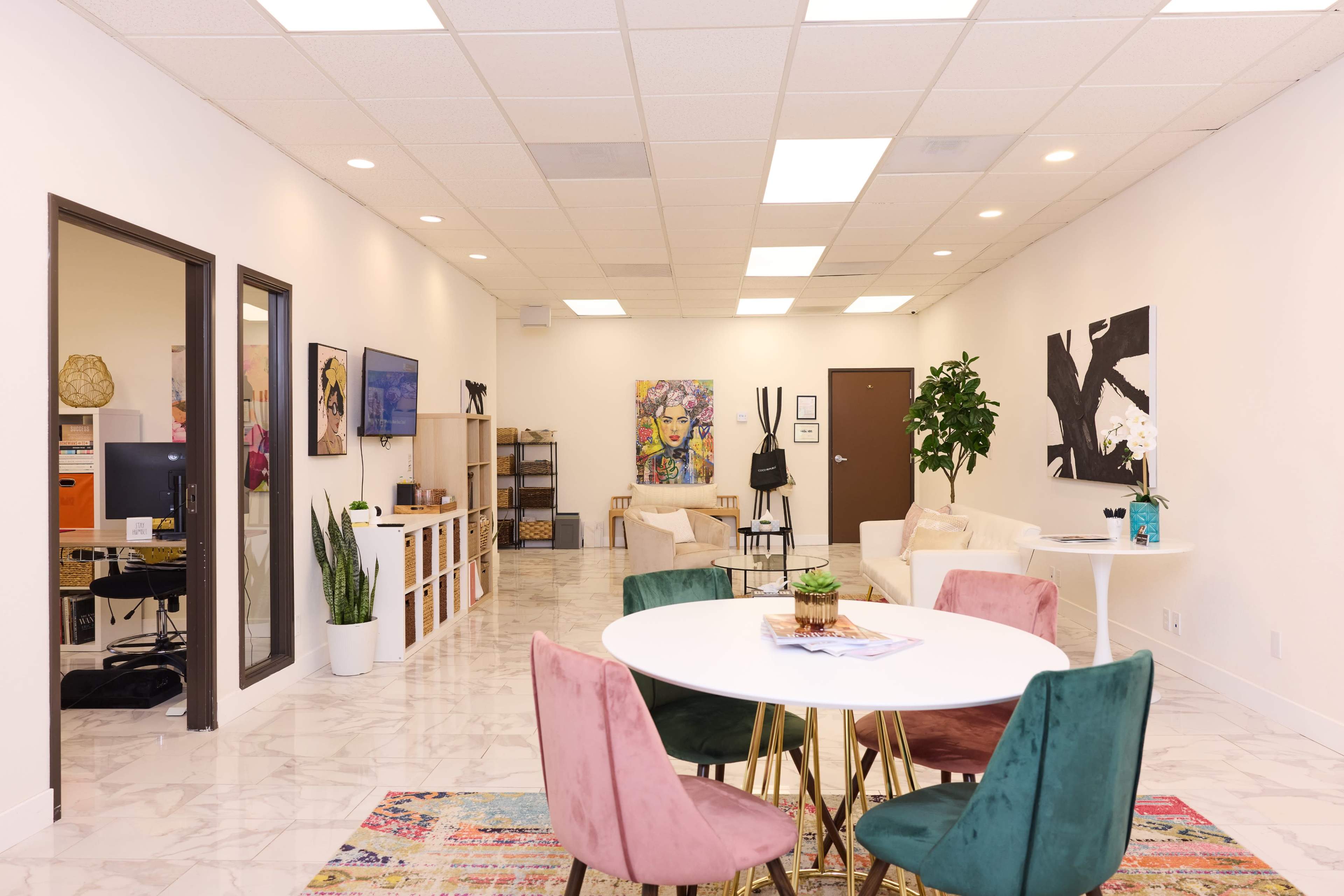A modern office space featuring a round table with colorful chairs, a seating area, artwork on the walls, and a reception area visible through an open doorway.