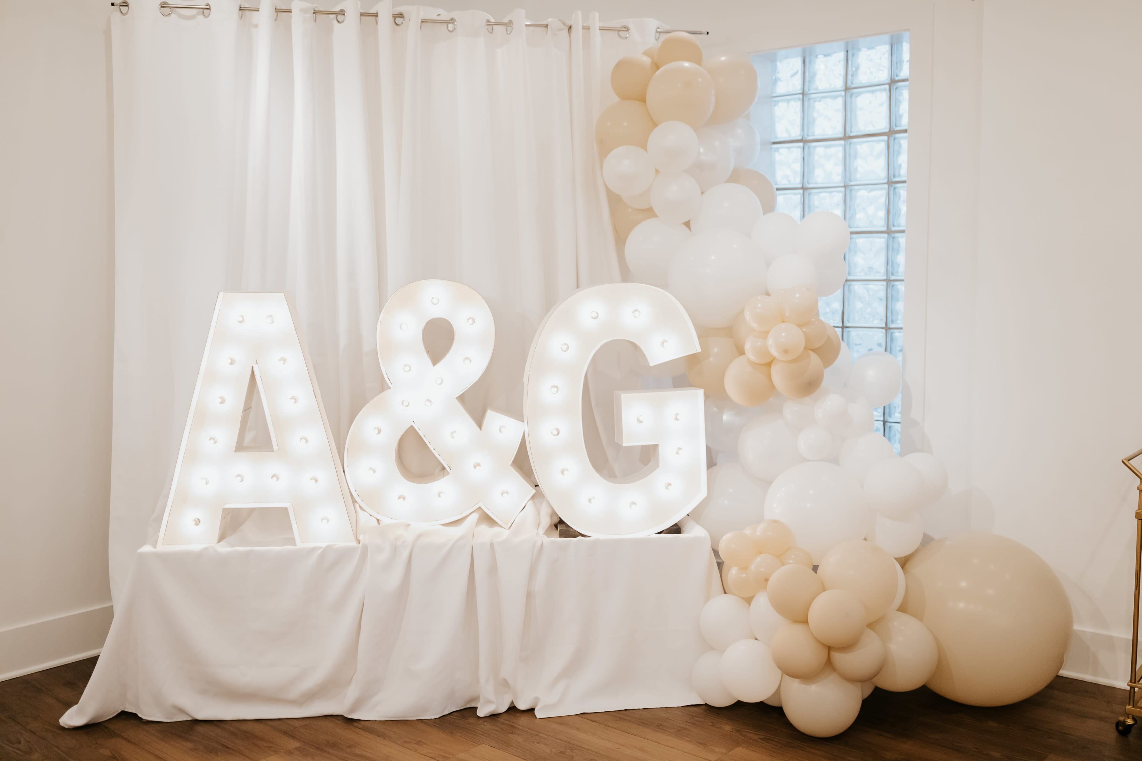 The image features illuminated letters "A" and "G" against a white curtain, accompanied by a decorative arrangement of balloons in neutral tones.
