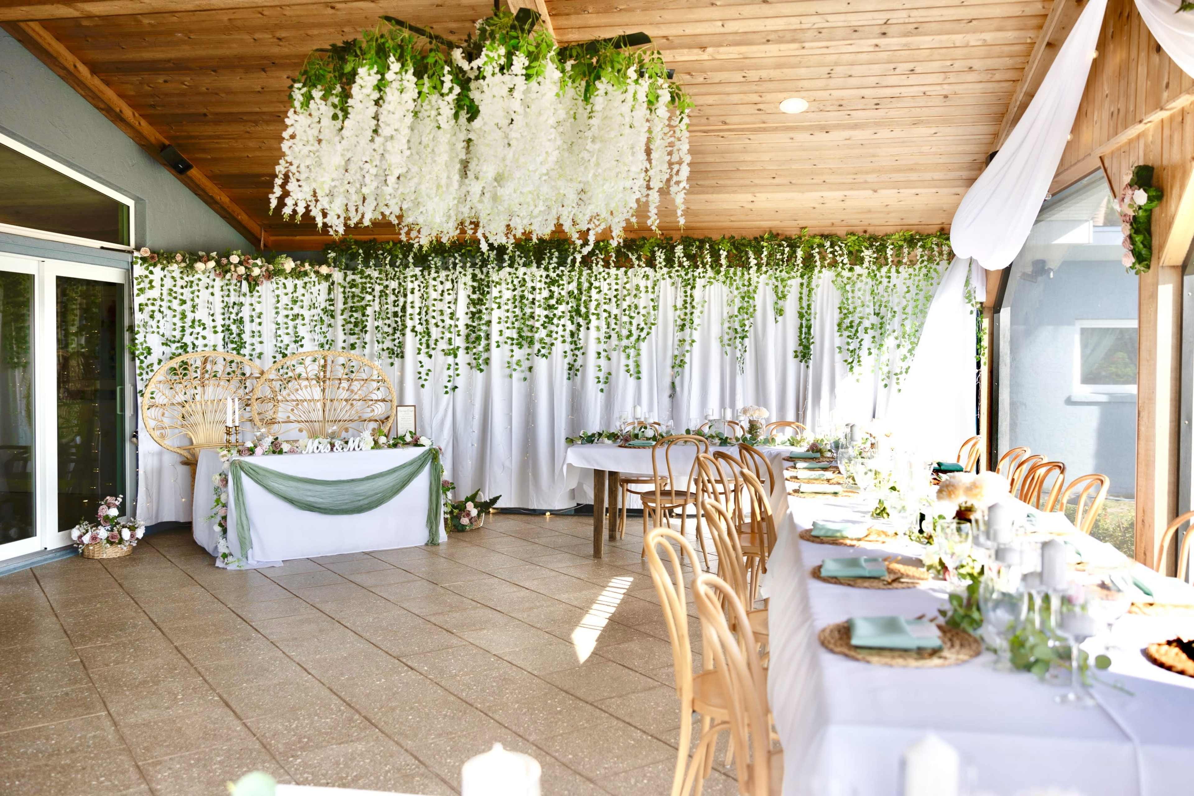 The image shows a decorated banquet hall with long tables set for an event, featuring greenery and floral arrangements hanging from the ceiling.
