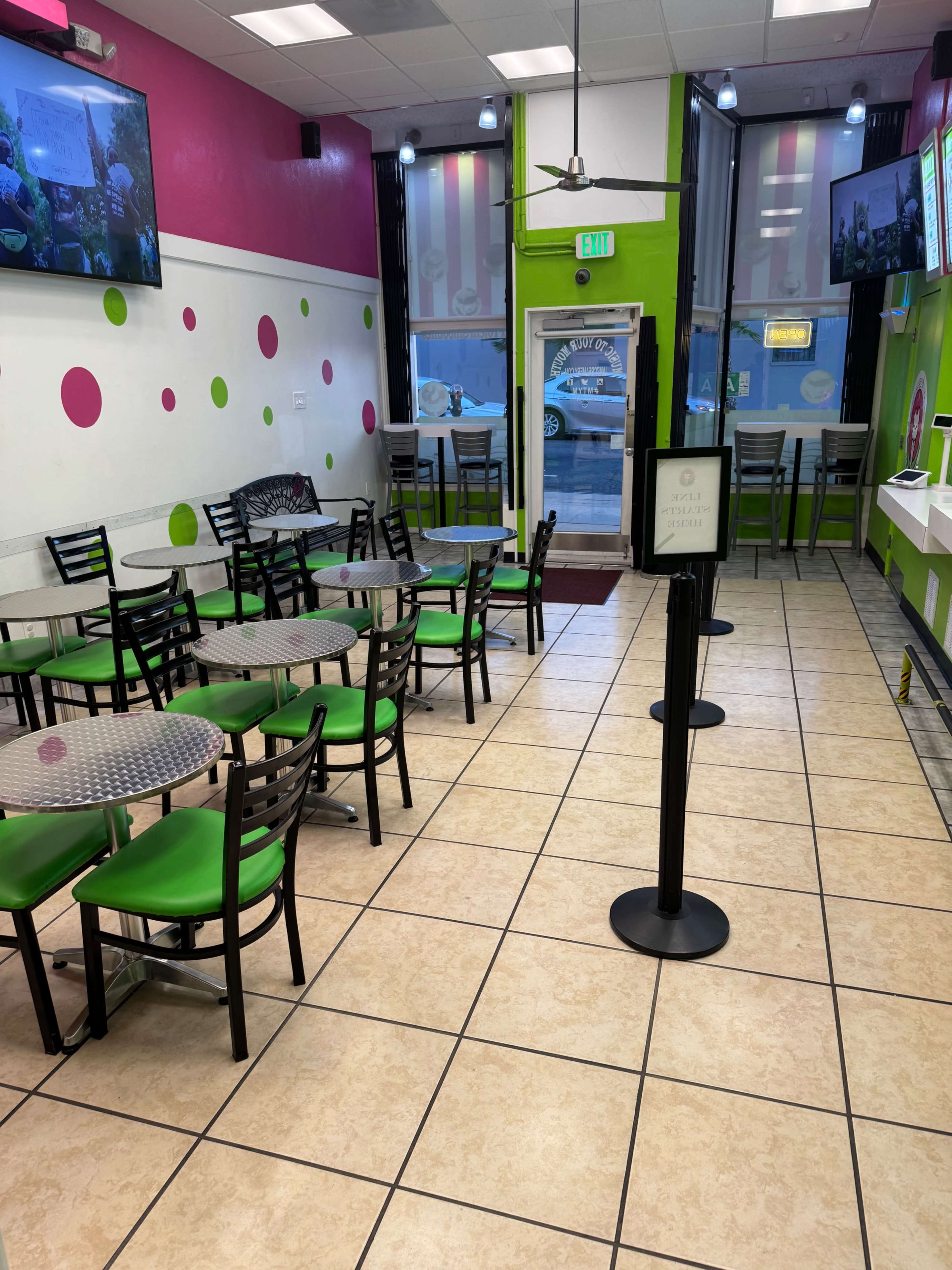 The image shows a brightly colored interior of a small restaurant with green and pink decor, featuring tables and chairs arranged neatly and a countertop at one end.
