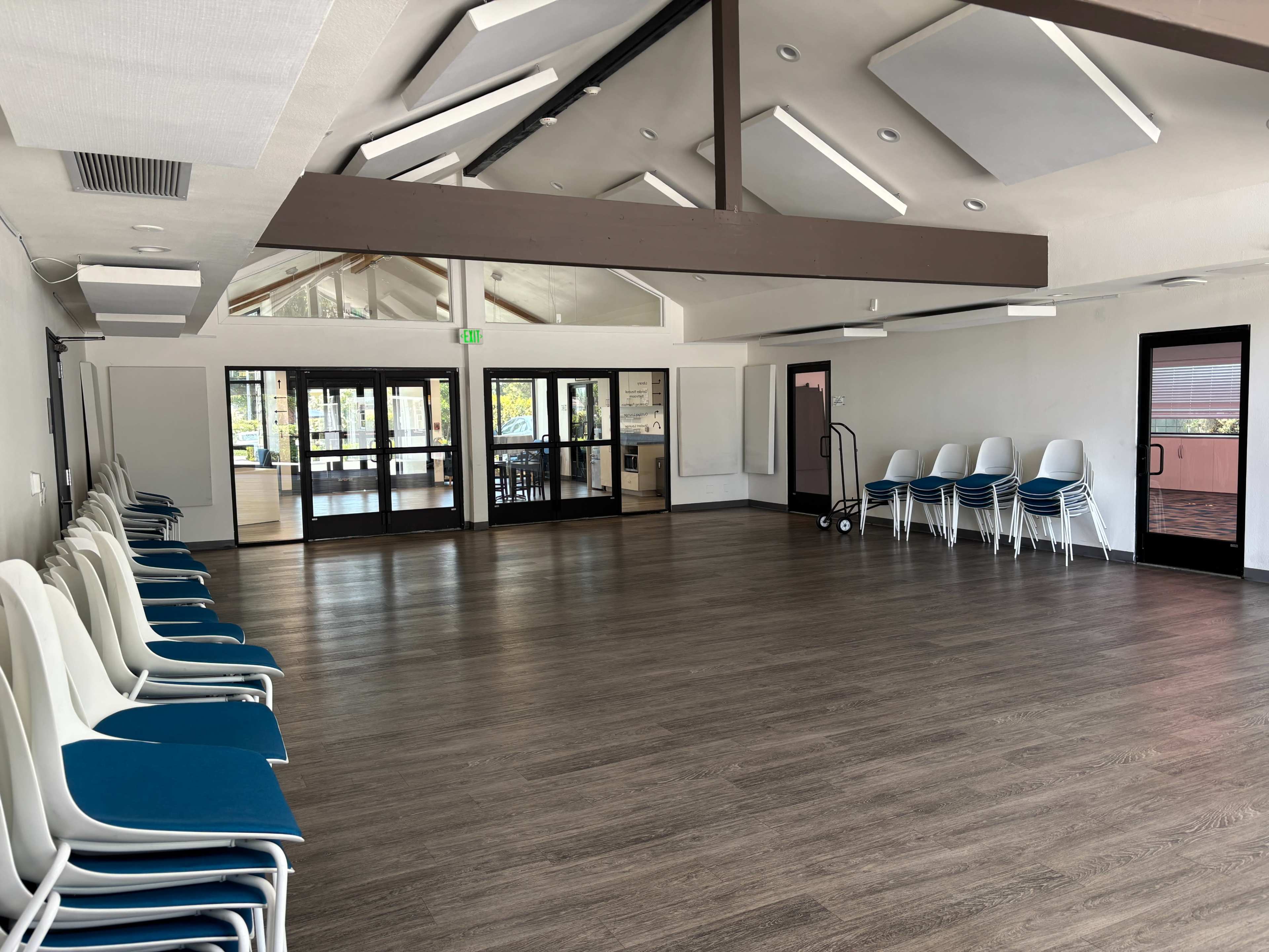 The image shows a spacious room with rows of white chairs against one wall, large windows allowing natural light, and a visible entrance area.