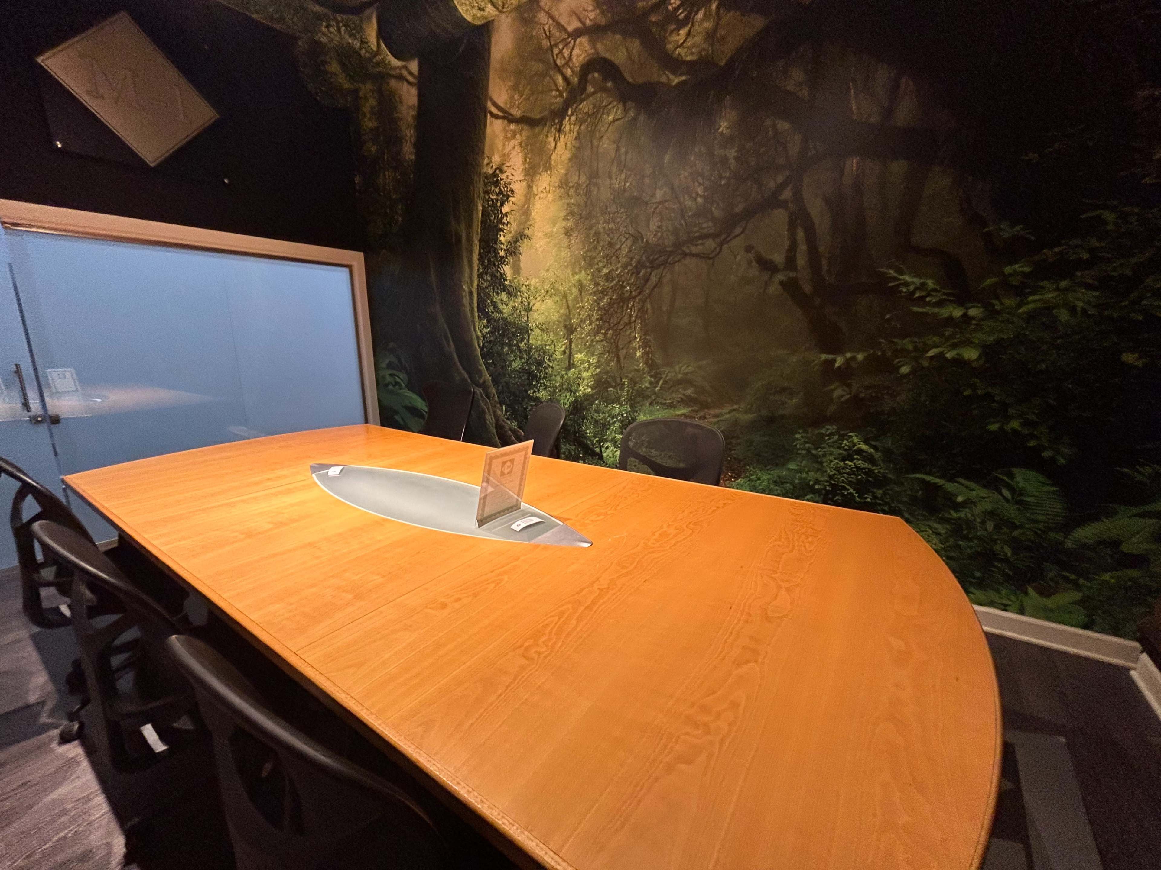 A conference room features a large wooden table surrounded by black chairs, set against a mural of a forest landscape.