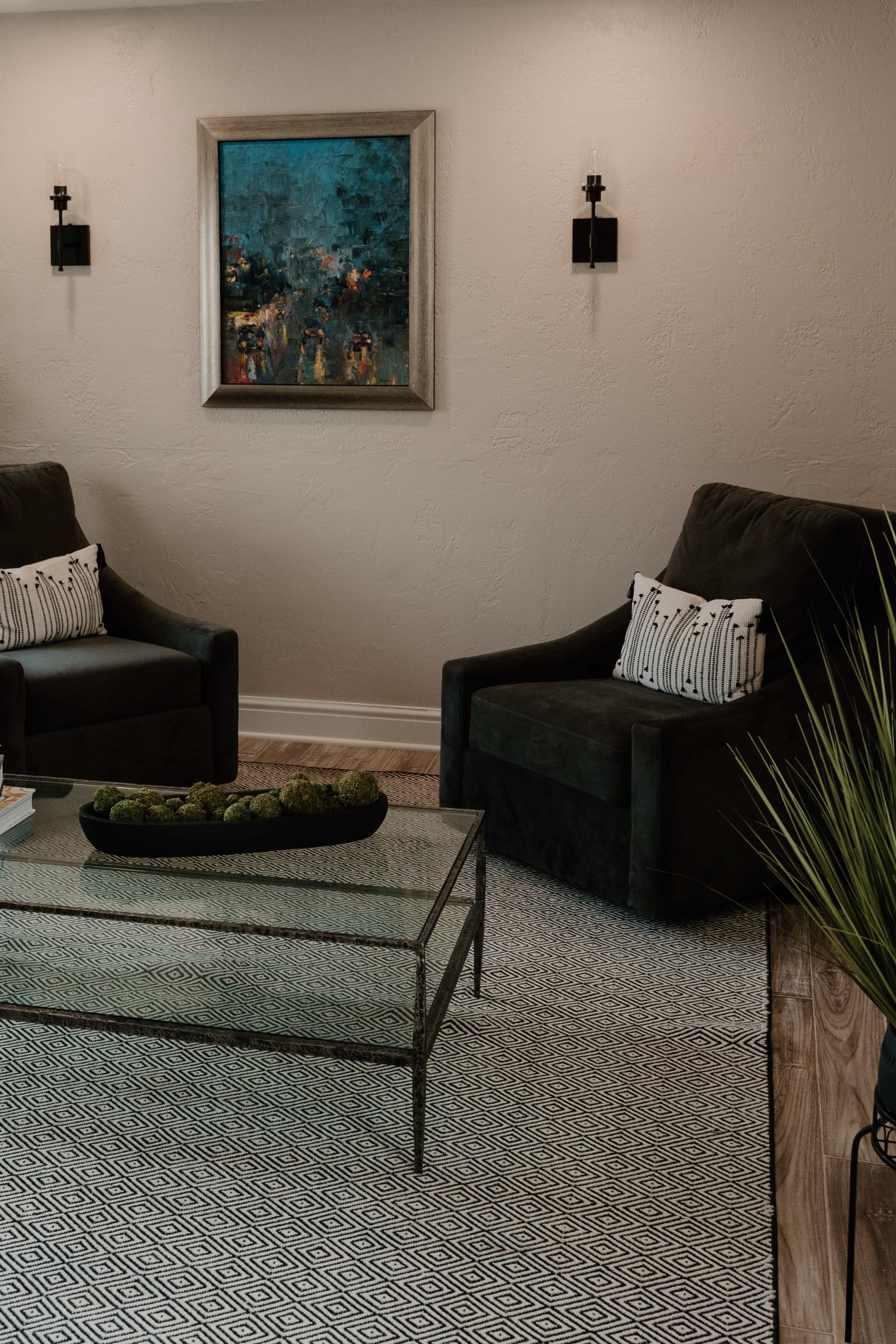 A living room with two armchairs flanking a glass coffee table on a patterned rug, and a colorful painting hanging on the wall.