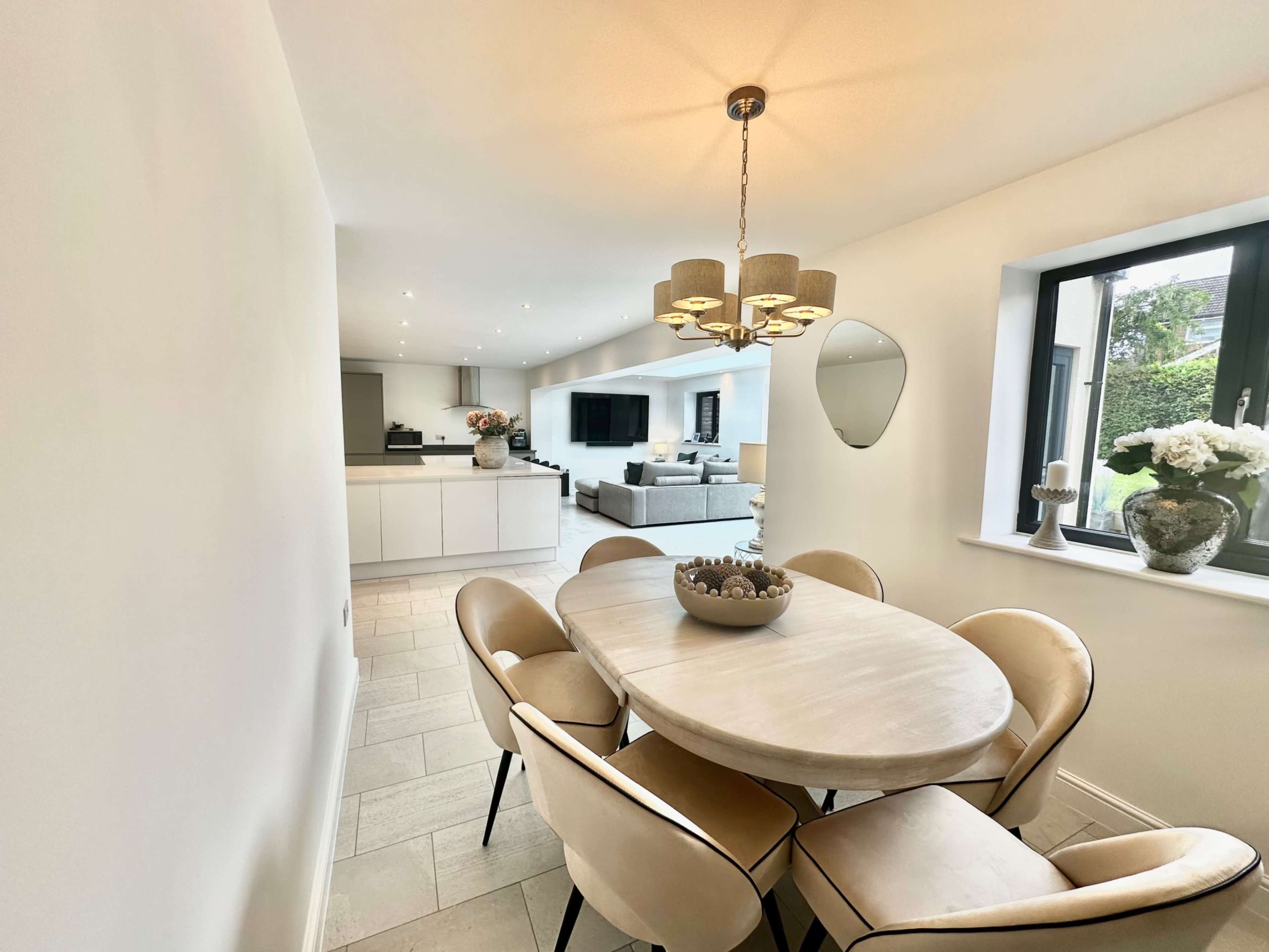 A dining area features a round table surrounded by upholstered chairs, with a chandelier overhead and large windows providing natural light.
