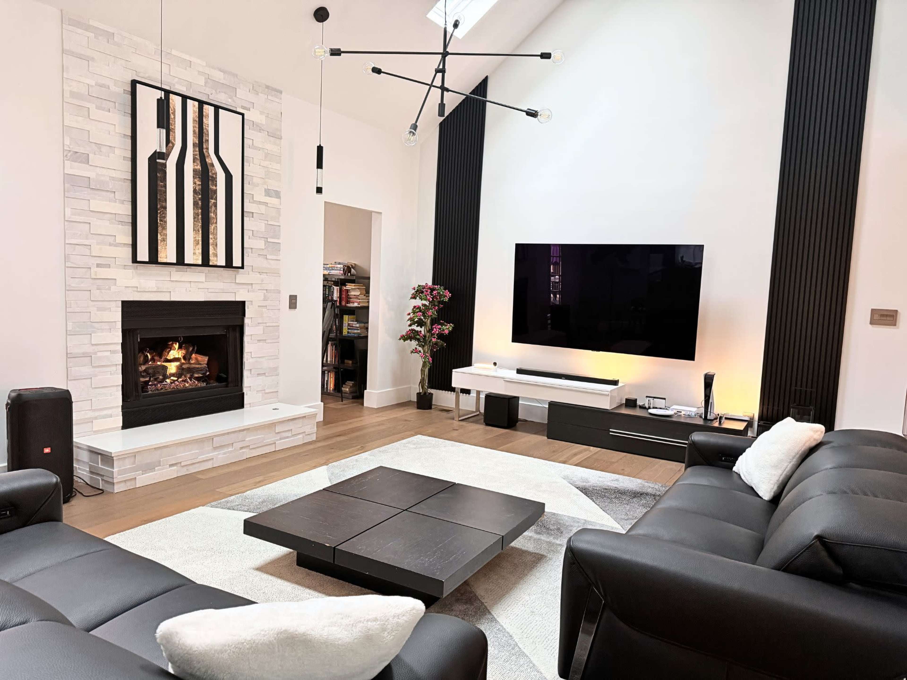 The image shows a modern living room with a stone fireplace, a large flat-screen TV mounted on the wall, and contemporary furniture arranged on a light-colored rug.