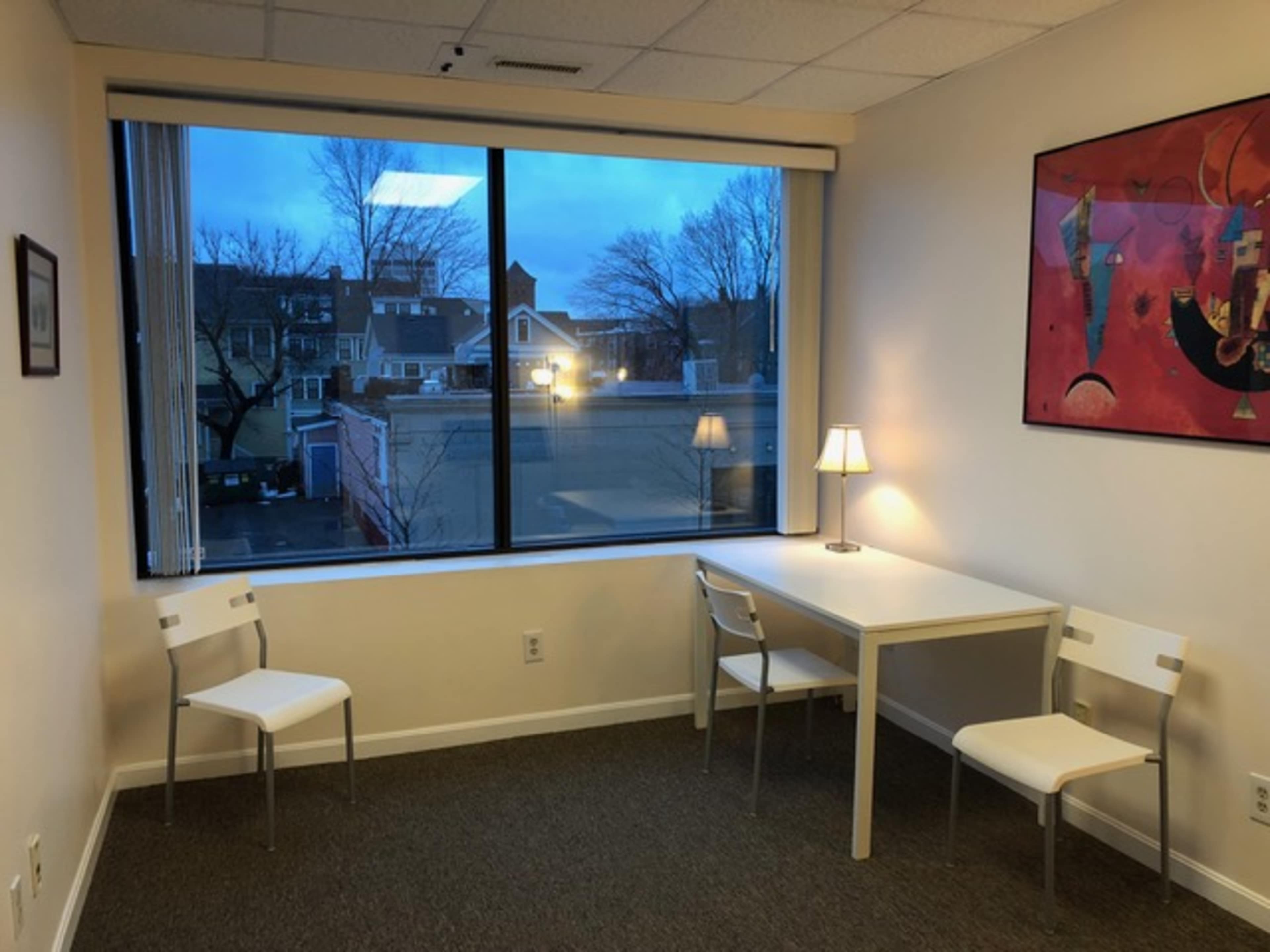 A small room with a white table and two chairs, featuring a window that overlooks a street with buildings and trees outside, illuminated by soft indoor lighting.