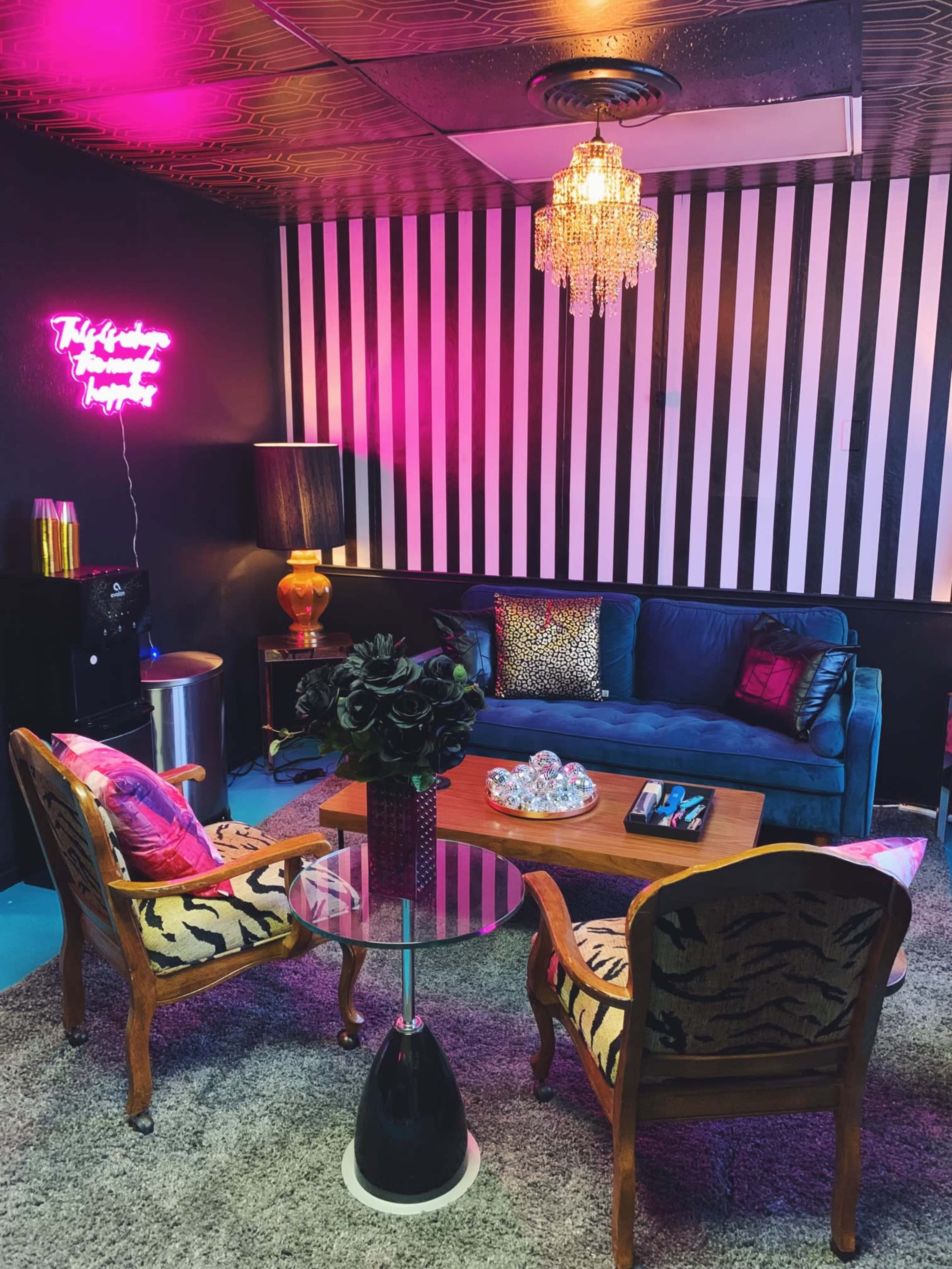 A cozy lounge area features a blue velvet sofa, two patterned chairs, a glass coffee table, and a neon sign on the wall, all under a chandelier.