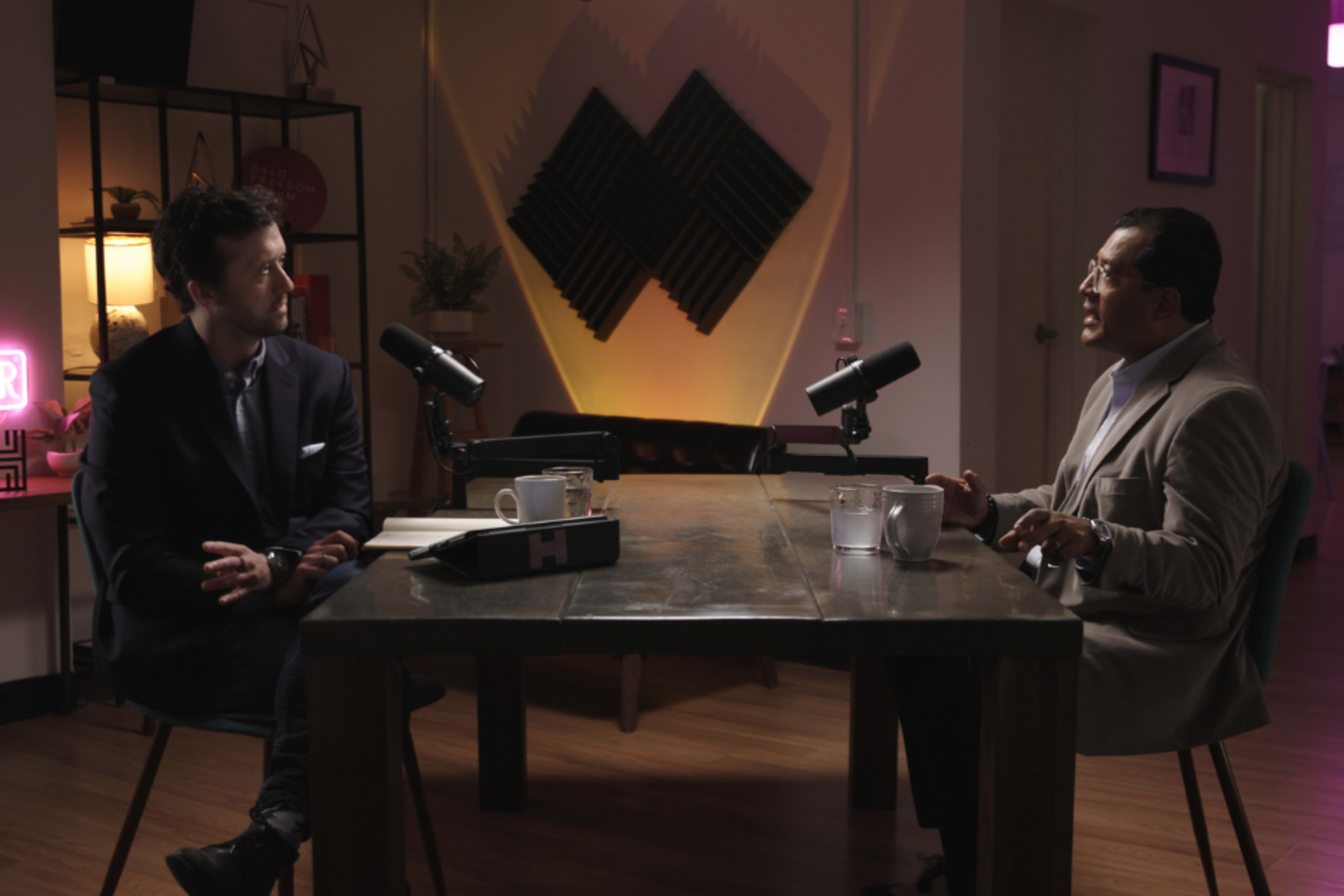 Two men are seated at a table in a dimly lit room, each facing a microphone, with notebooks and cups positioned on the table between them.