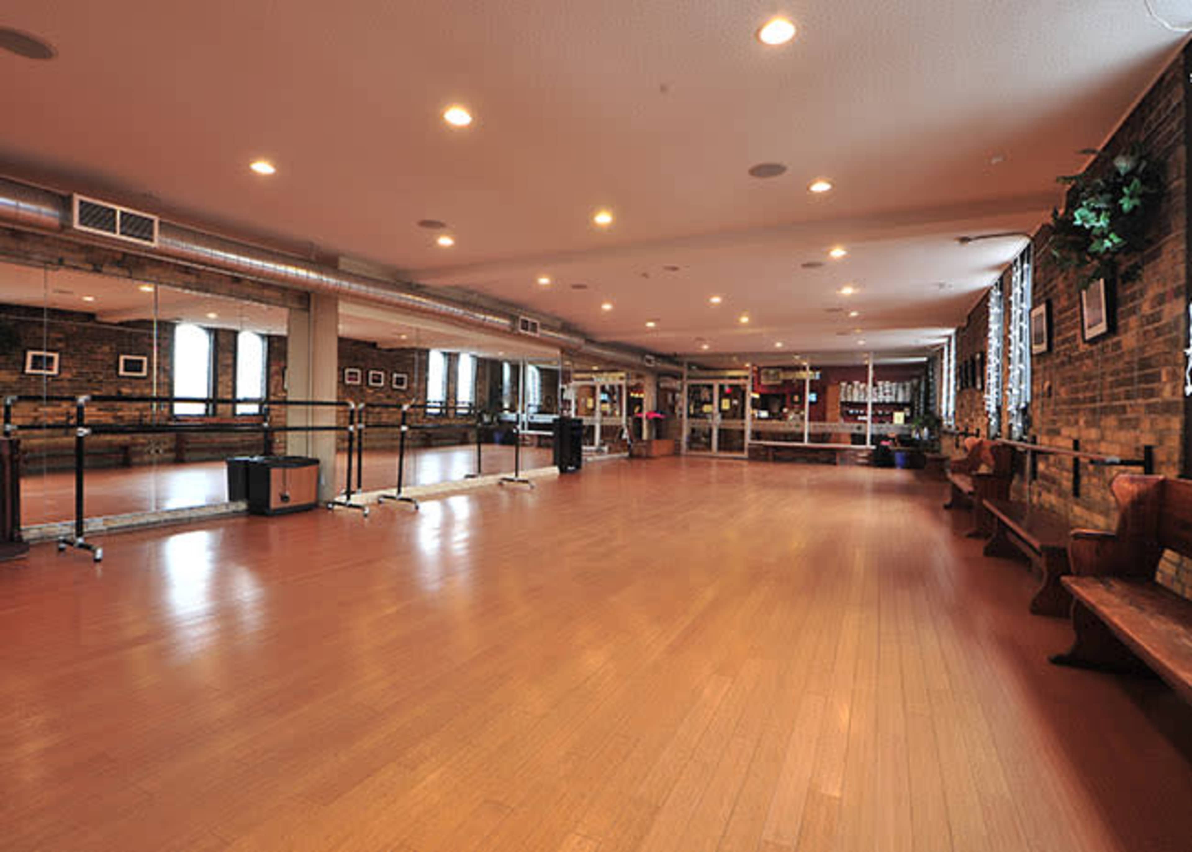 The image shows a spacious dance studio with wooden floors, mirrored walls, and benches along one side.