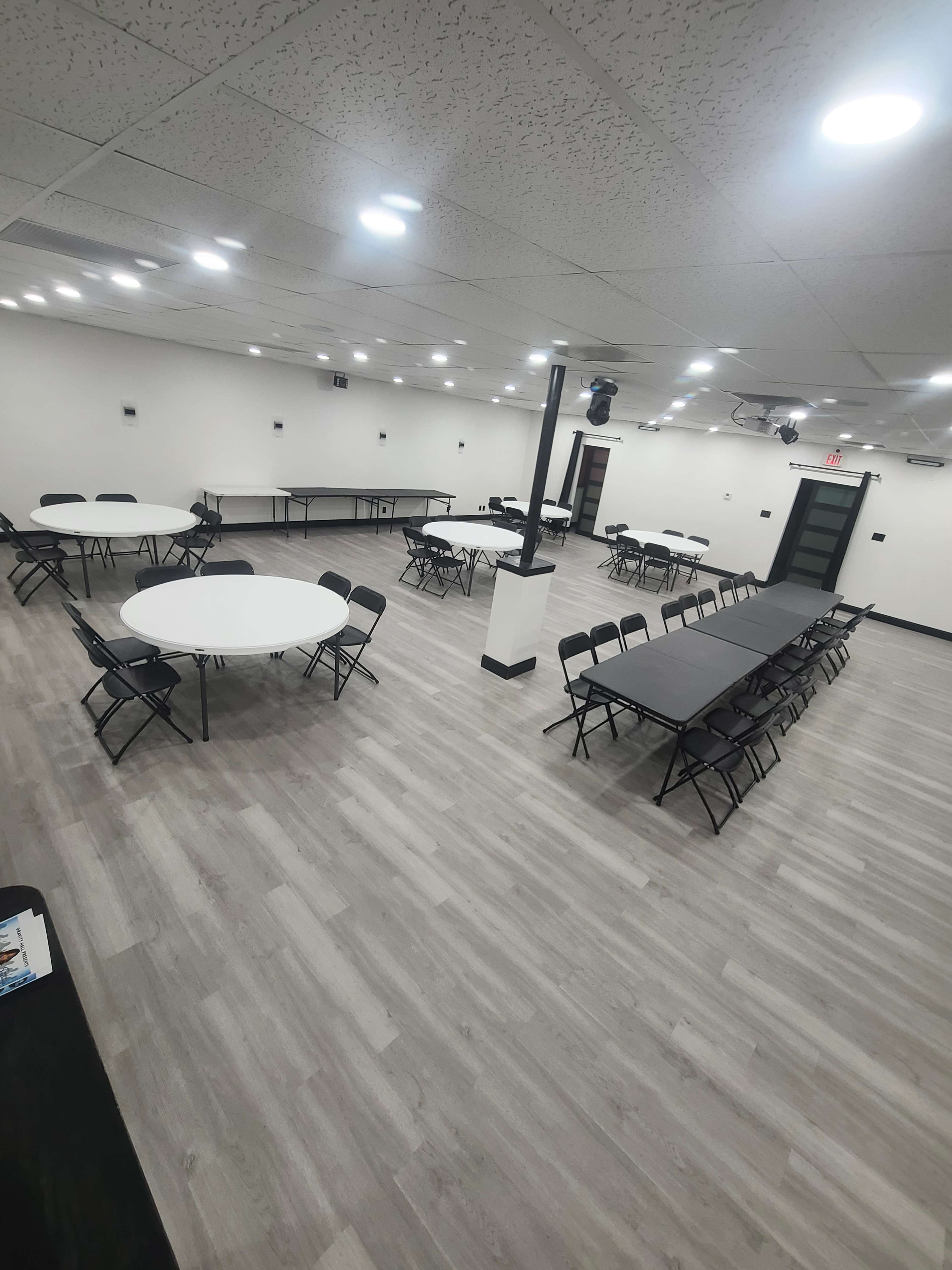 The image shows a spacious meeting room with various tables set up, including round and rectangular configurations, and black chairs arranged around them.