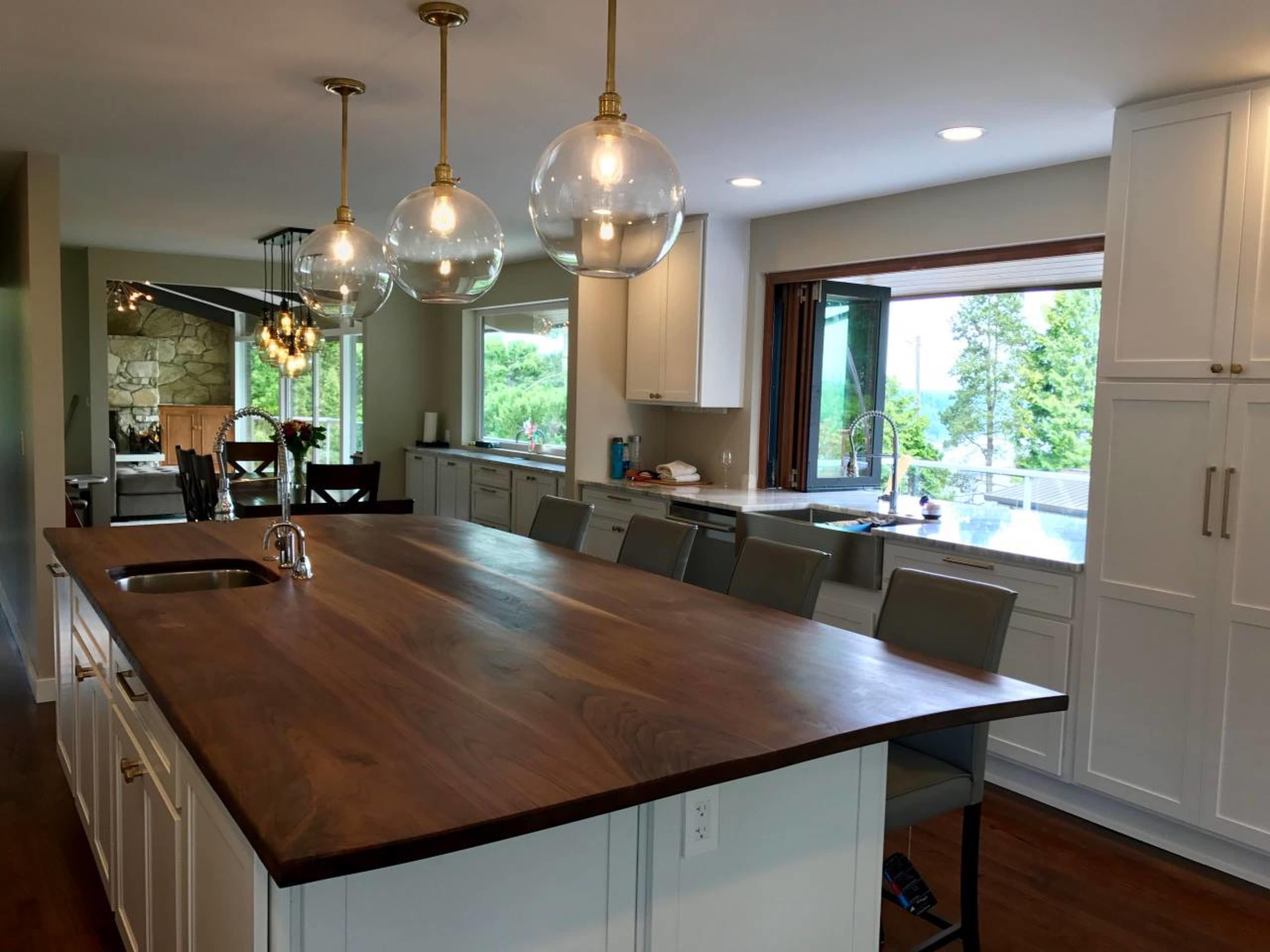 A modern kitchen with a large wooden island, pendant lights overhead, and a view of trees through a window.