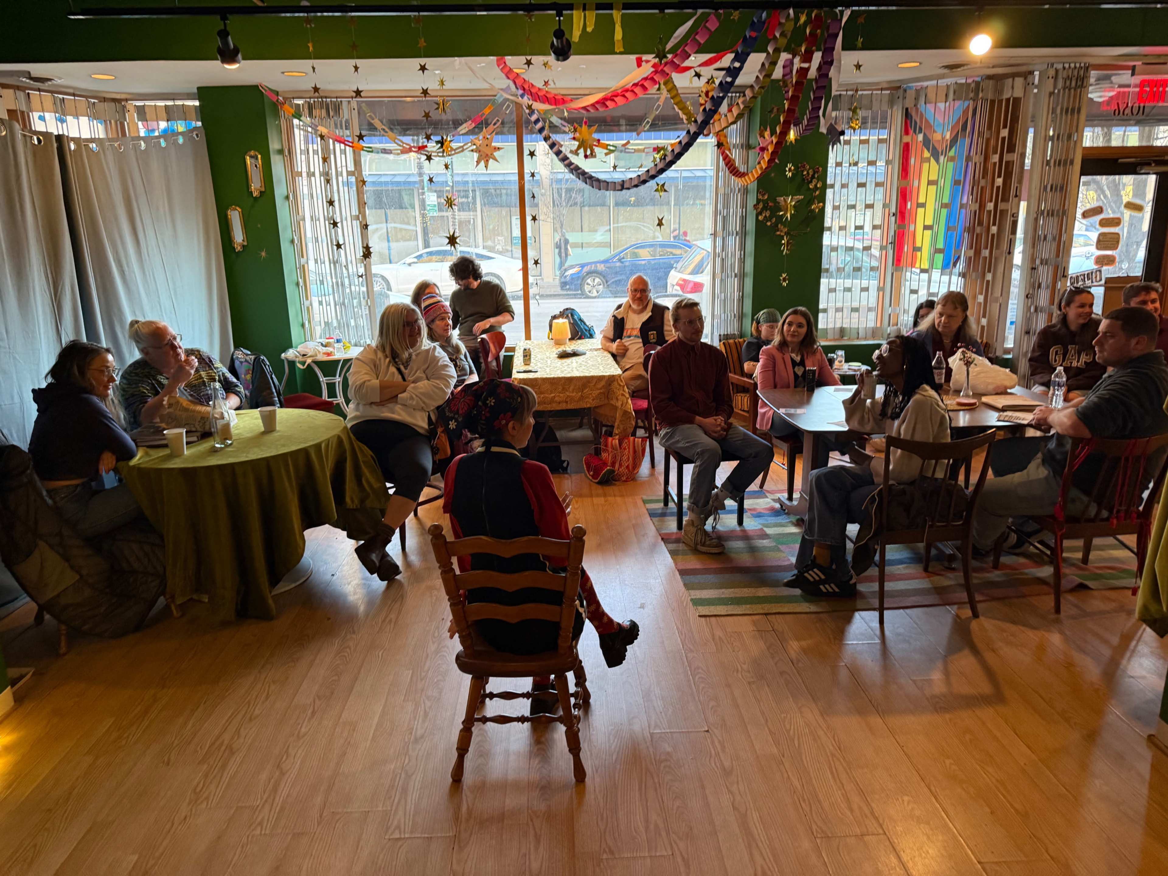 A group of people sits at tables in a colorful cafe, some engaged in conversation while others watch a speaker in front.