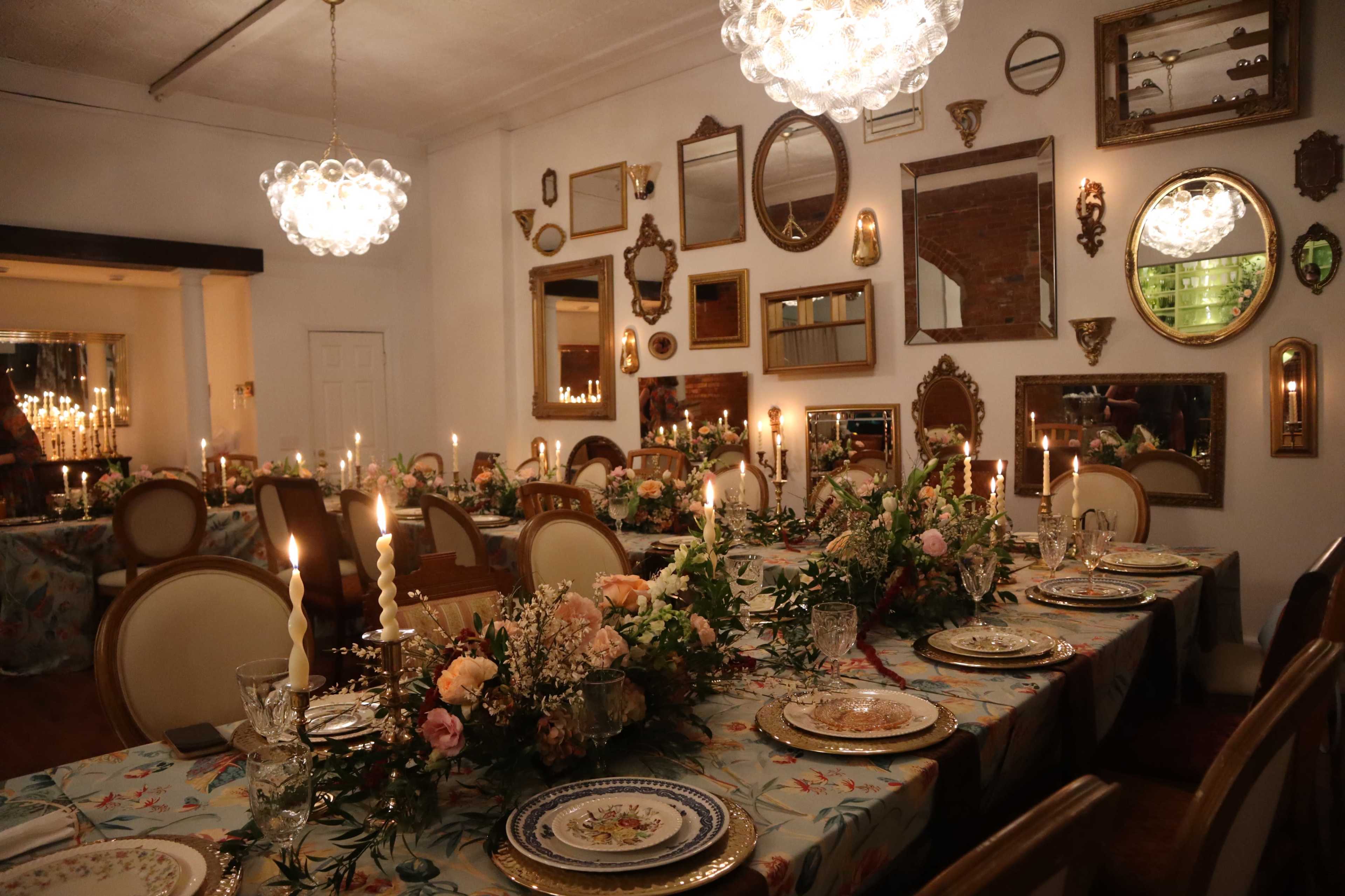A dining room is elegantly set with tables adorned with floral arrangements, candles, and fine china, surrounded by a variety of framed mirrors on the walls.