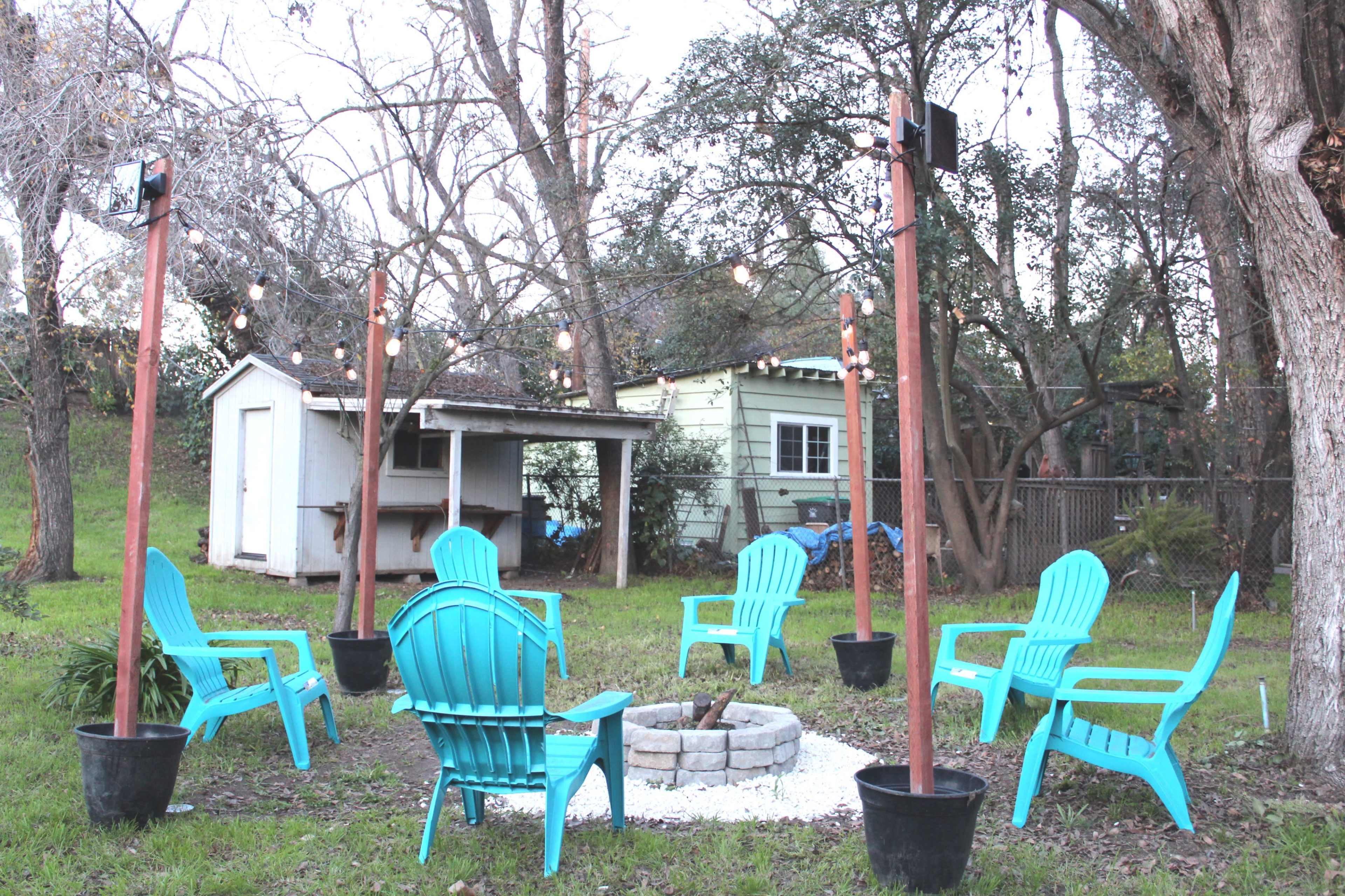 A circular gathering space with blue adirondack chairs surrounds a stone fire pit, with string lights overhead and a shed in the background.