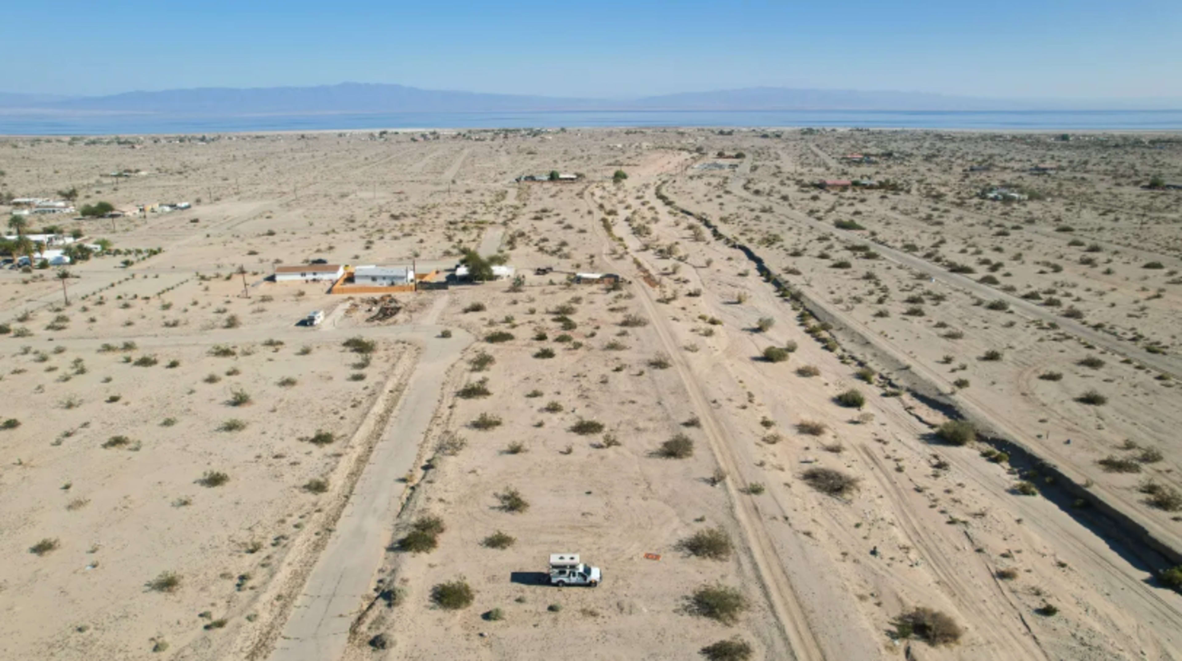 A barren desert landscape with sparse vegetation, scattered buildings, and an RV parked on a dirt road.