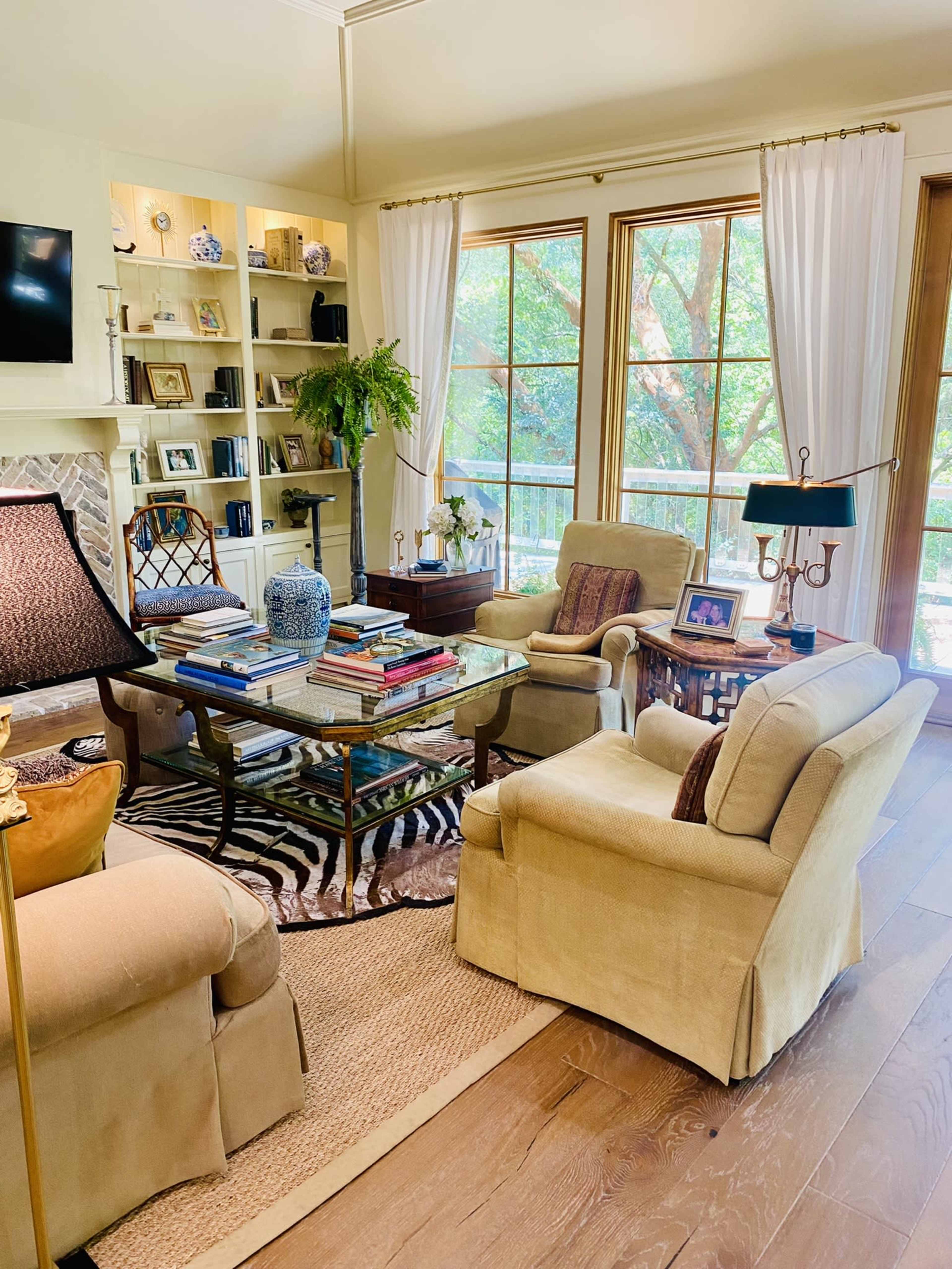 A cozy living room features two beige chairs, a glass coffee table with stacked books, and large windows providing a view of greenery.