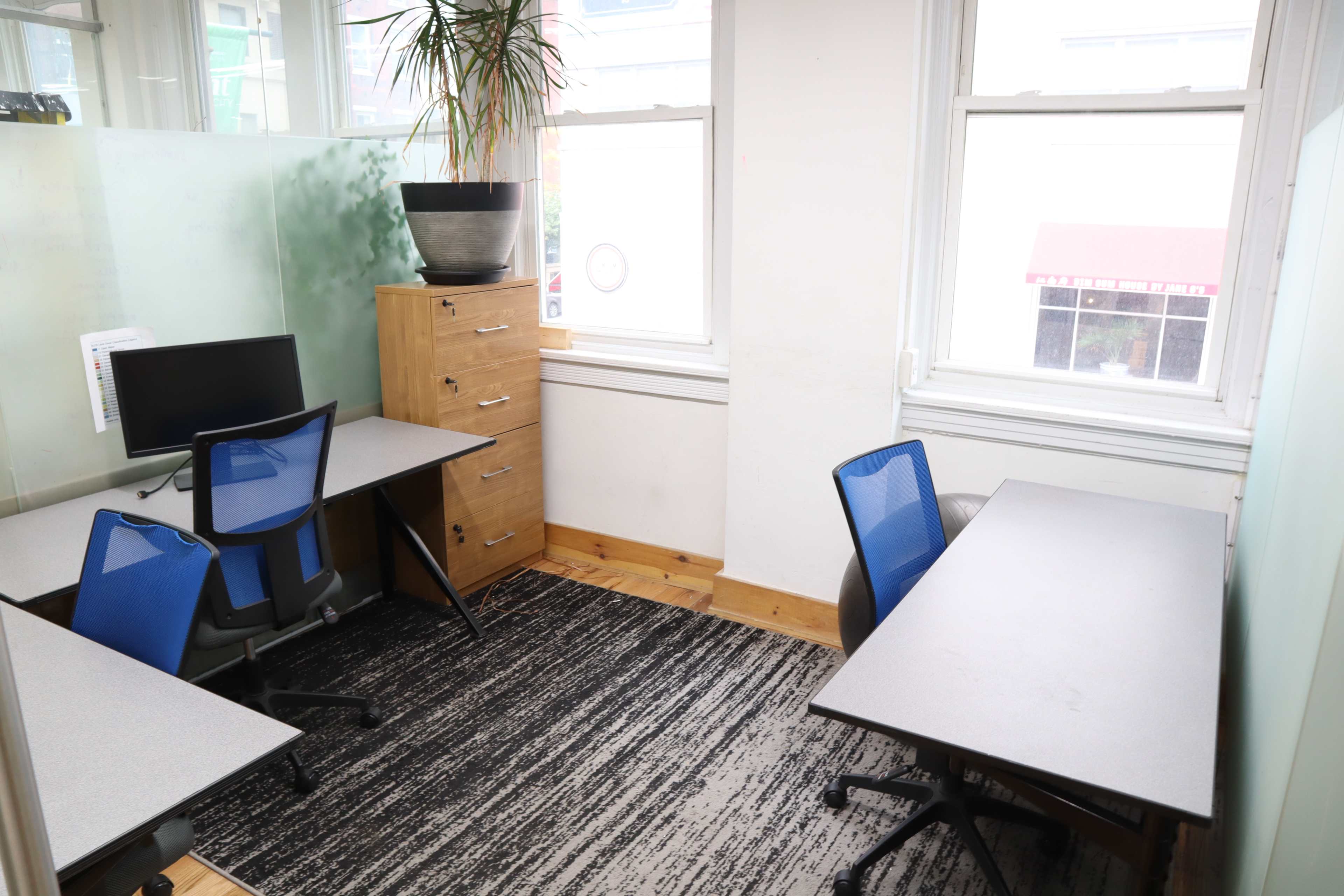 The image shows a small office space with two desks, a computer, a filing cabinet, and large windows allowing natural light to enter.