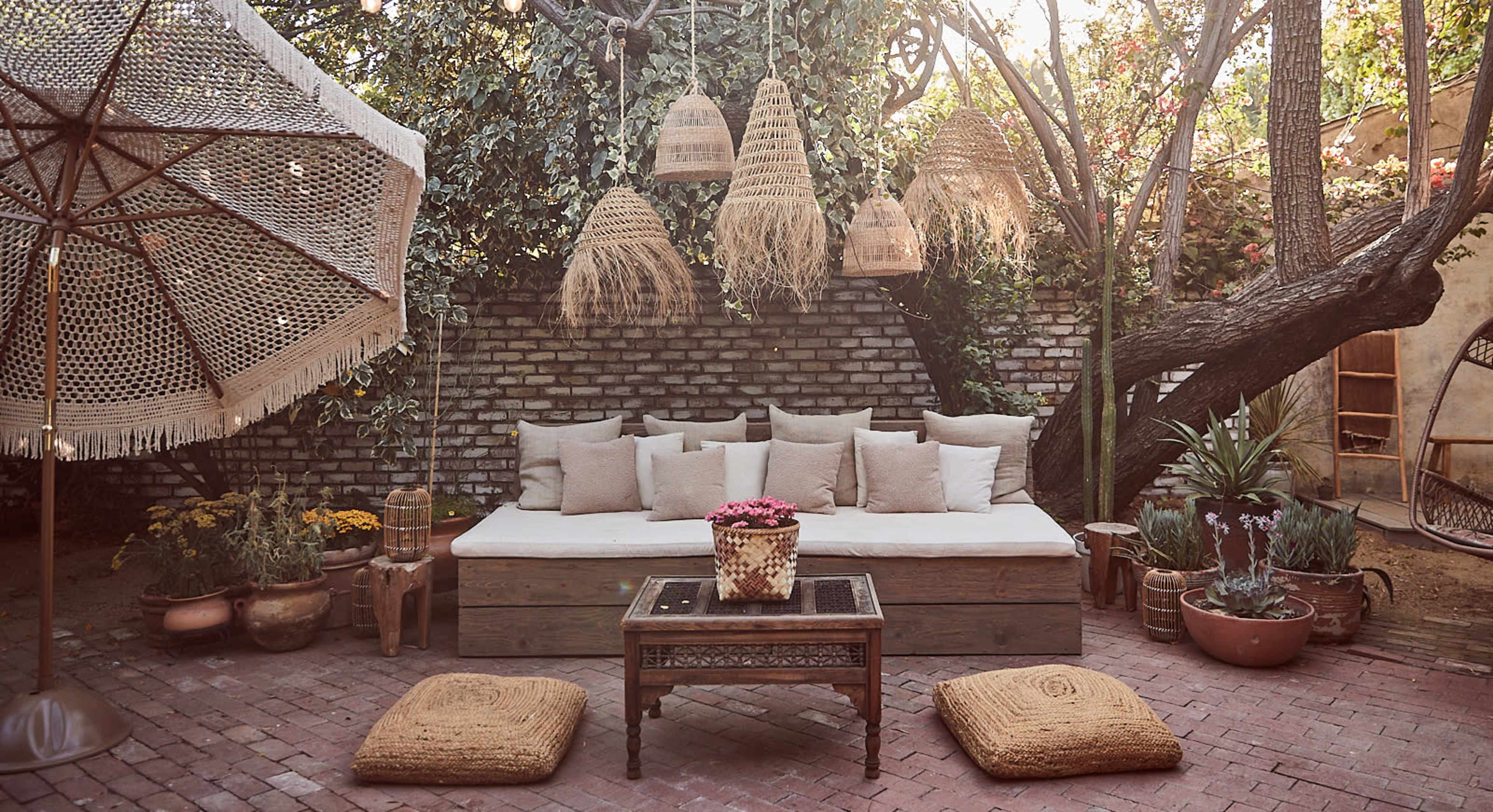 A cozy outdoor seating area with a large wooden sofa, decorative hanging lights, a coffee table, and plants, surrounded by brick walls and a large umbrella.