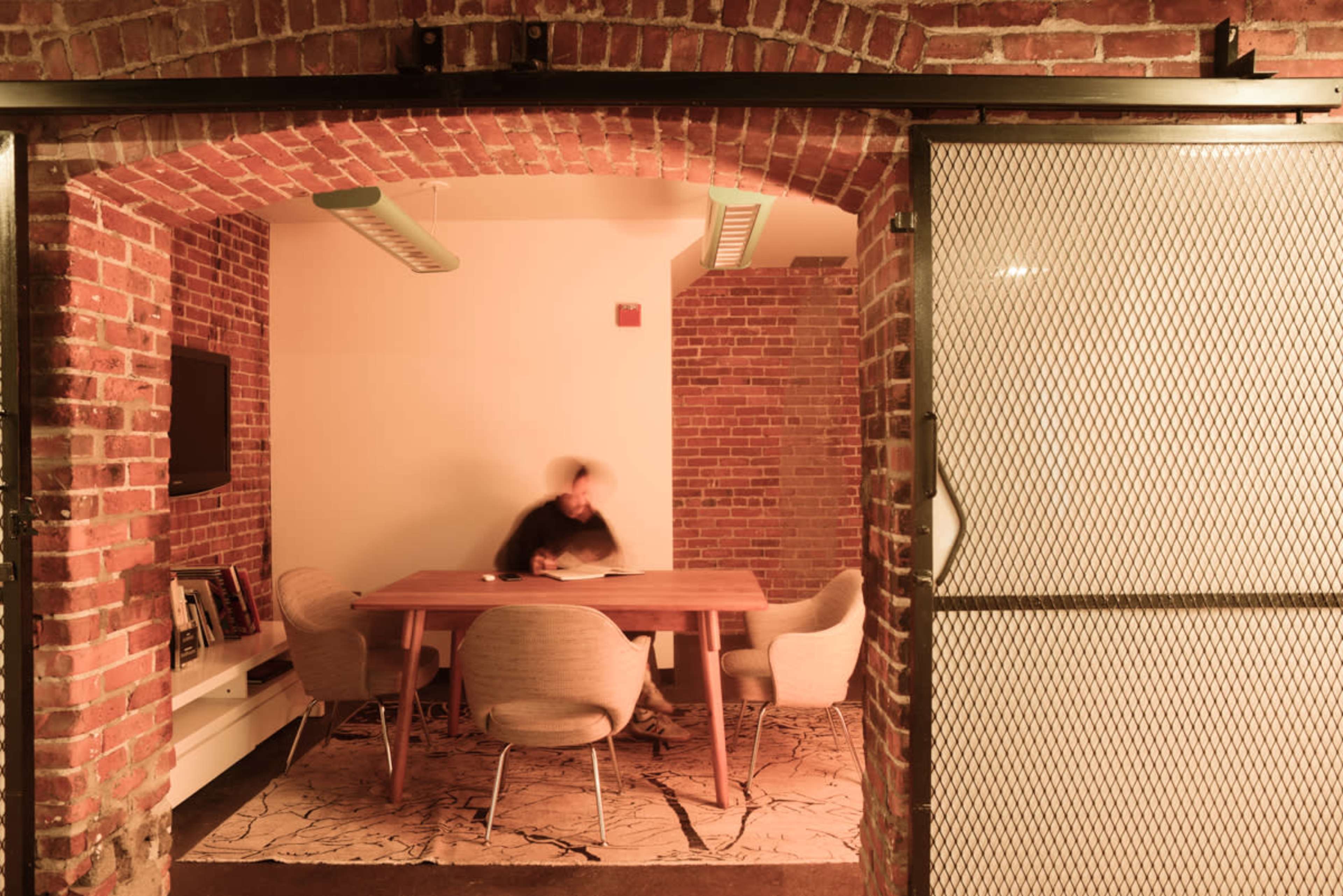 A meeting room with a large table and four chairs, characterized by exposed brick walls and ambient lighting.