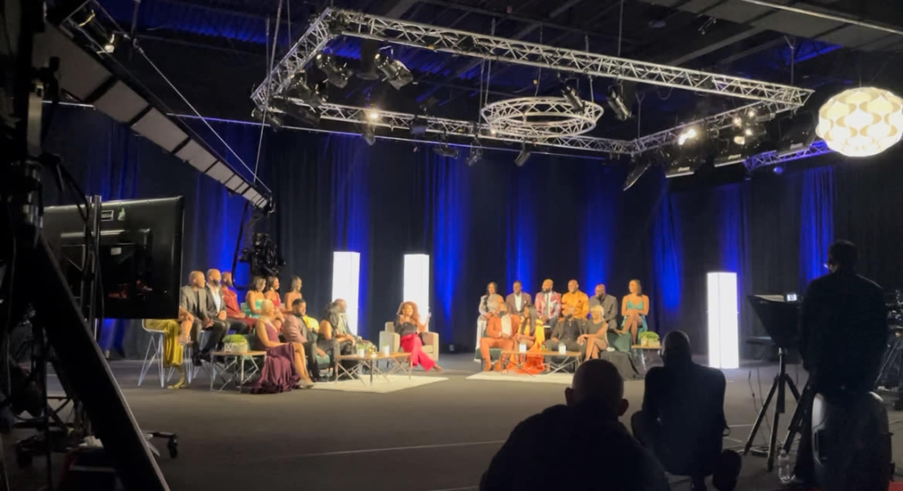 A diverse group of people is seated in a brightly lit studio arranged for a discussion or presentation, surrounded by professional lighting equipment.