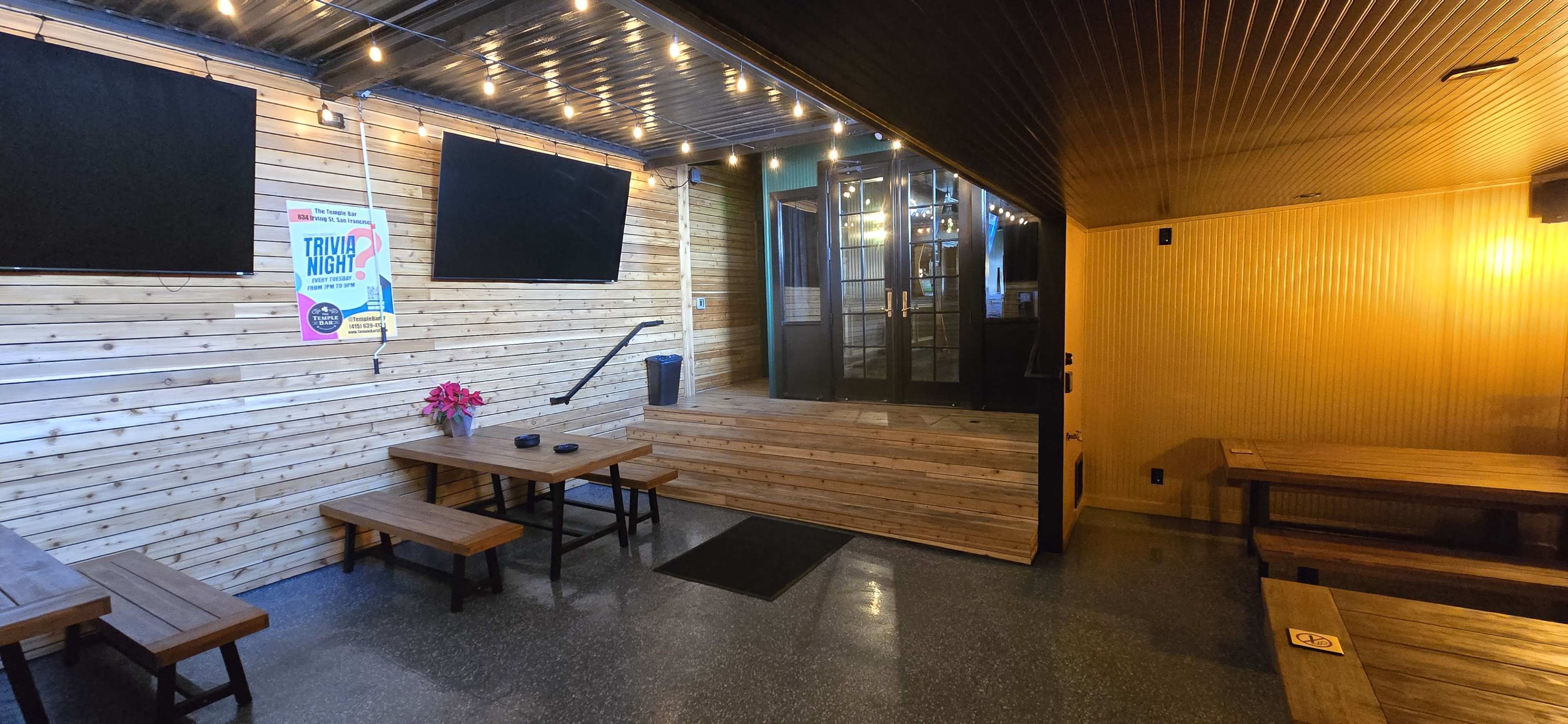 The image shows a cozy indoor space with wooden tables and benches, featuring a staircase leading to a glass door entrance, surrounded by wooden paneling and string lights.