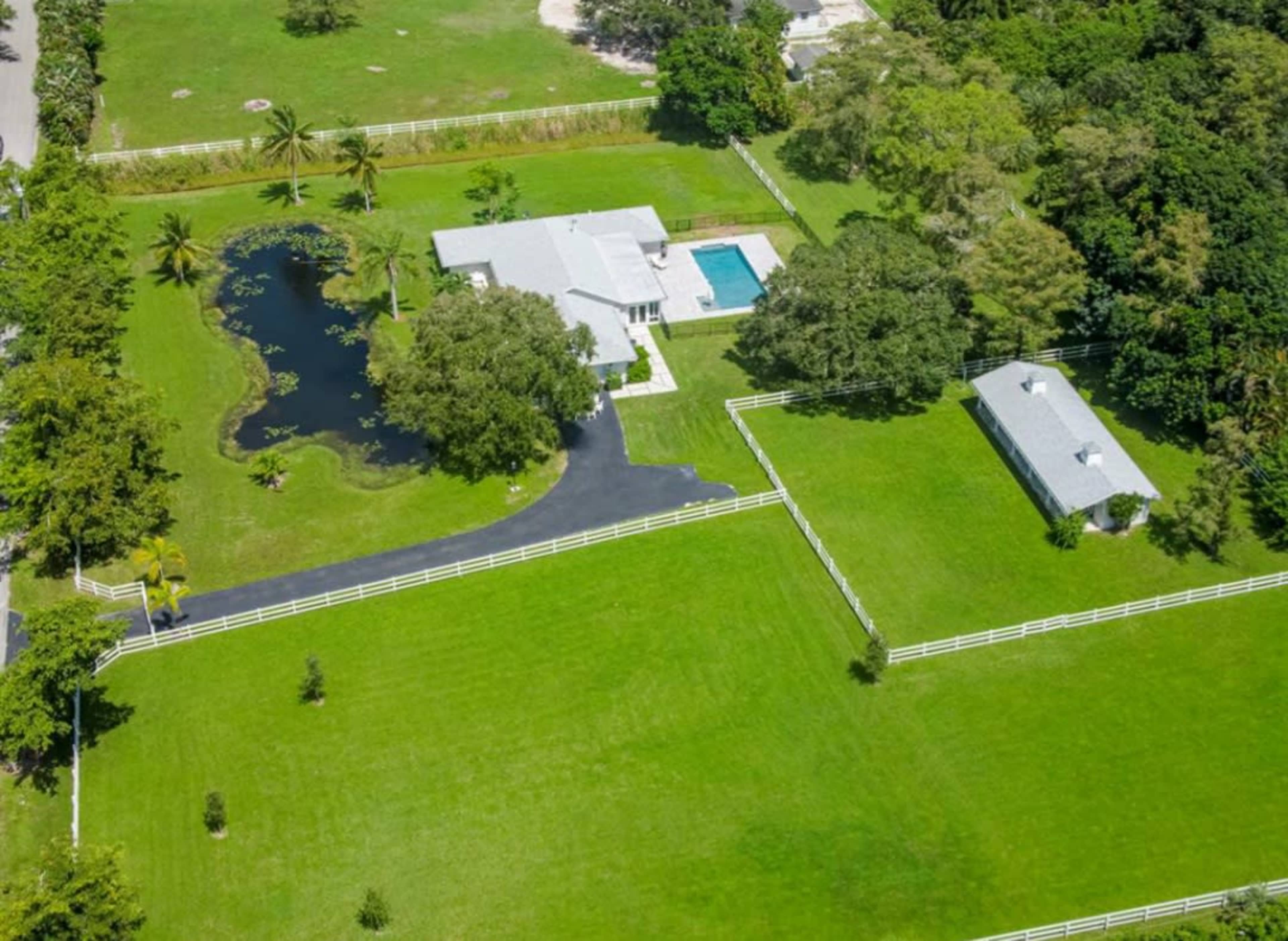 An aerial view shows a house with a pool, a pond, and fenced green fields surrounded by trees.
