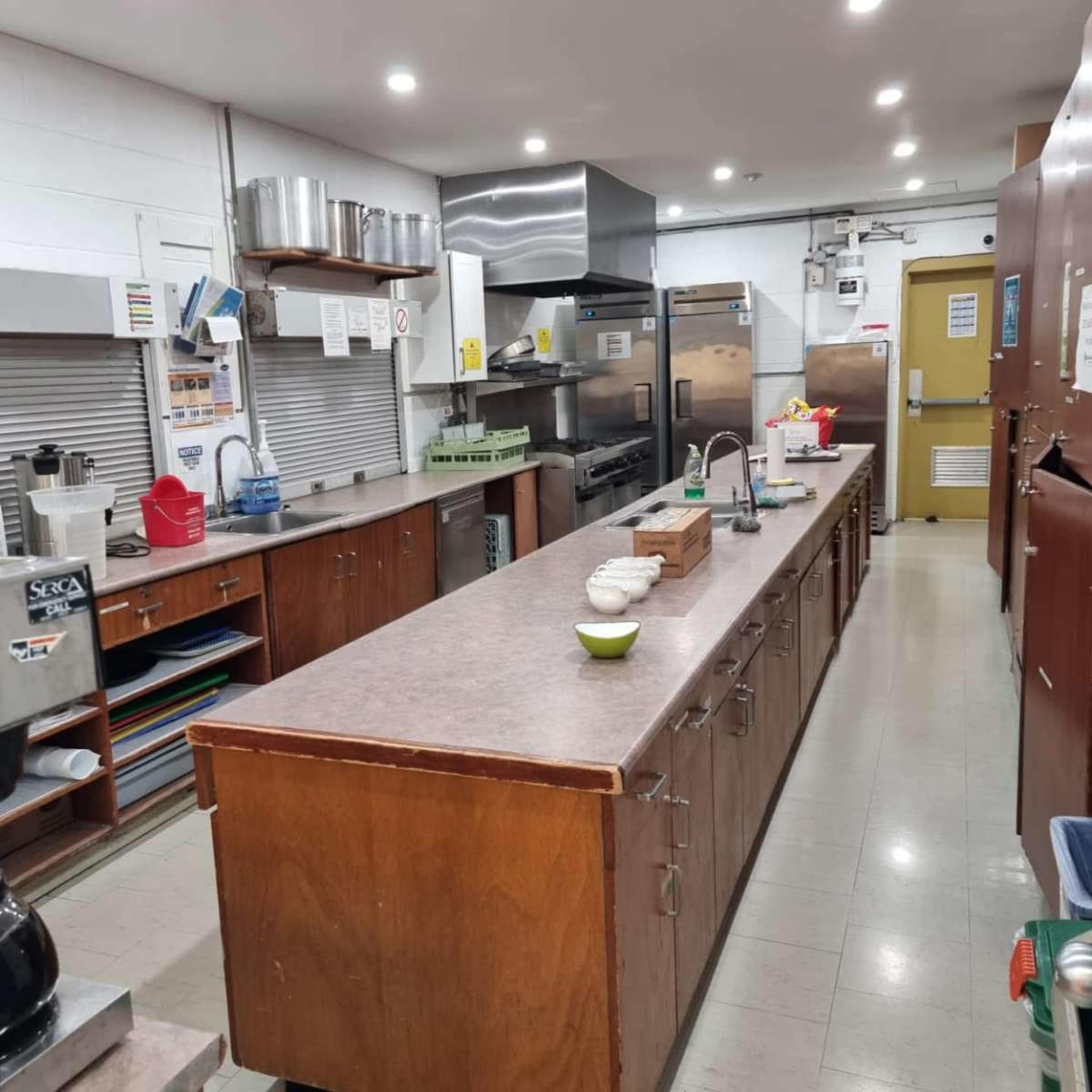 The image shows a clean and organized kitchen with a long countertop, various appliances, and shelving along the walls.