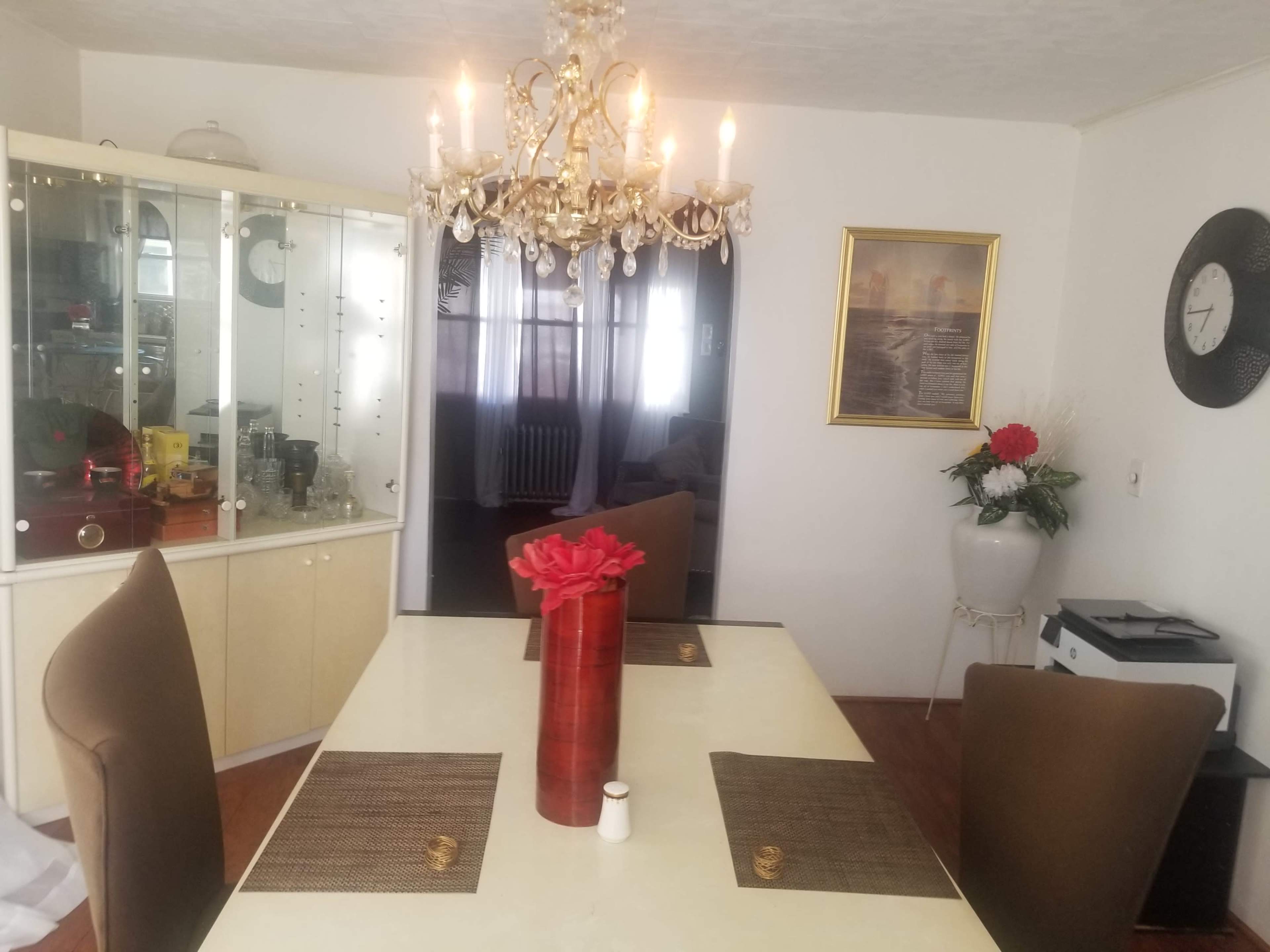 A dining room with a chandelier above a table set with placemats, surrounded by four chairs, a glass display cabinet, and a decorative plant.