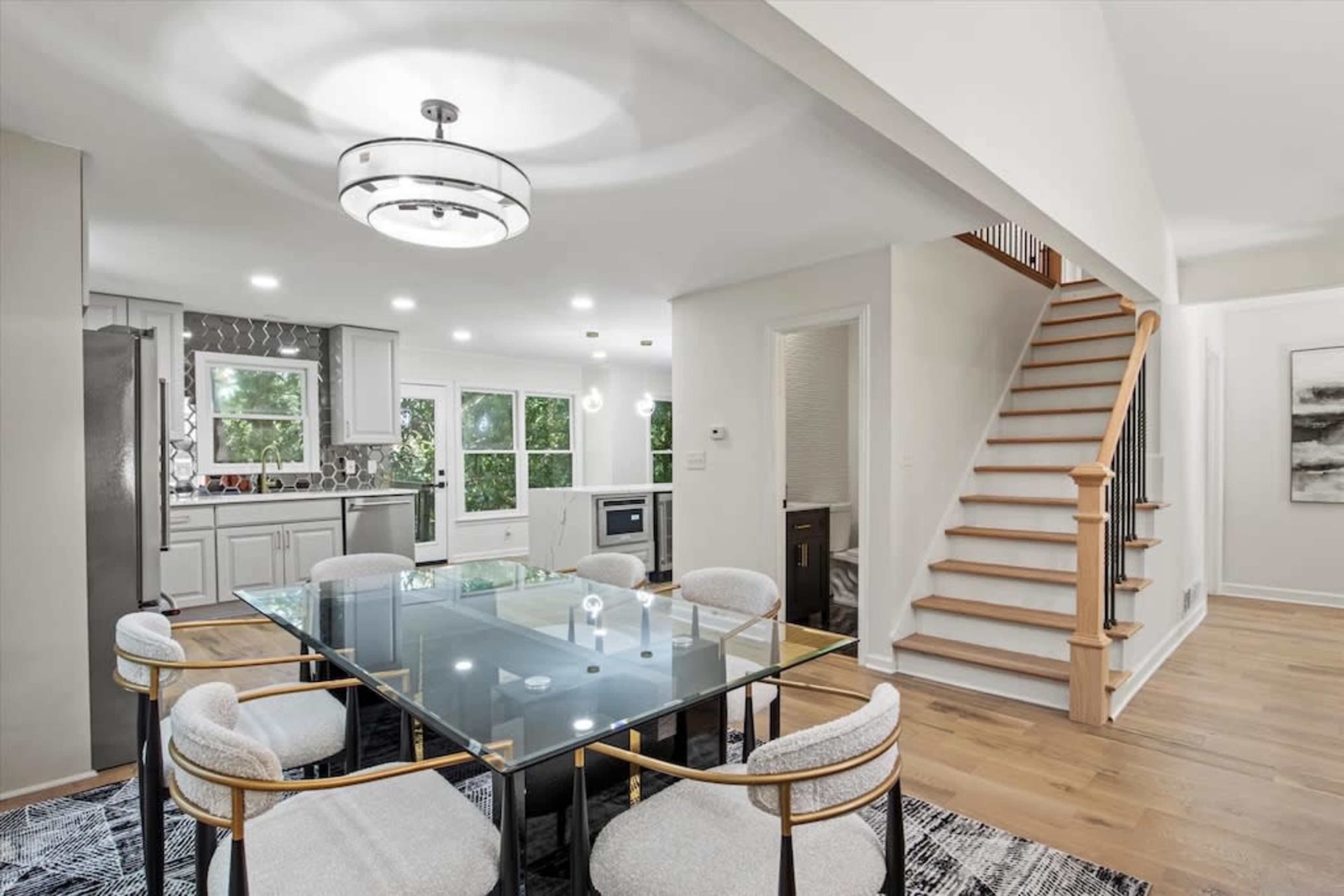 The image shows a modern dining area featuring a glass table surrounded by upholstered chairs, adjacent to a kitchen and a wooden staircase.
