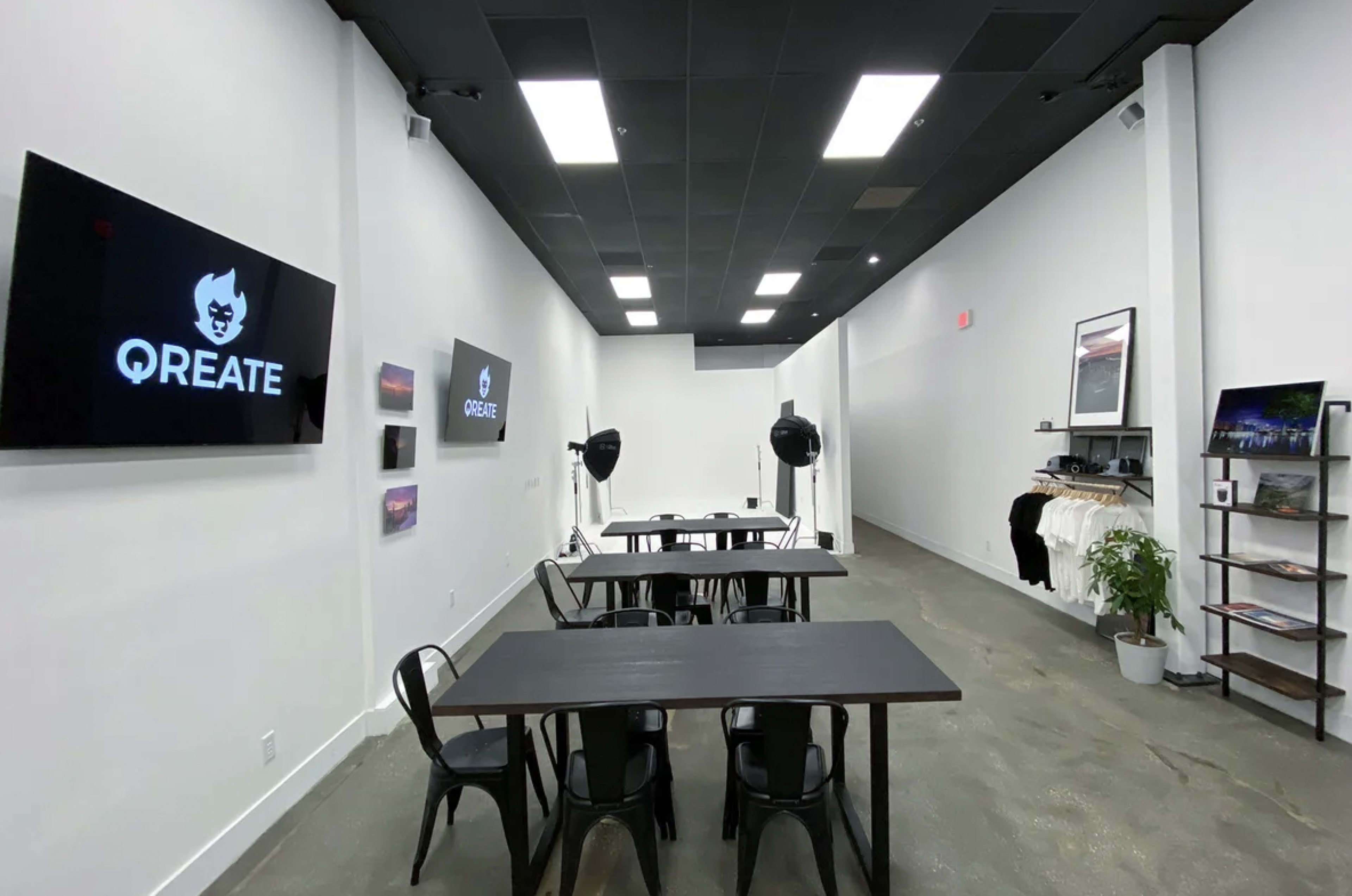 A modern, minimalistic workspace with several tables, wall-mounted screens displaying the QREATE logo, and various studio equipment in an empty room.