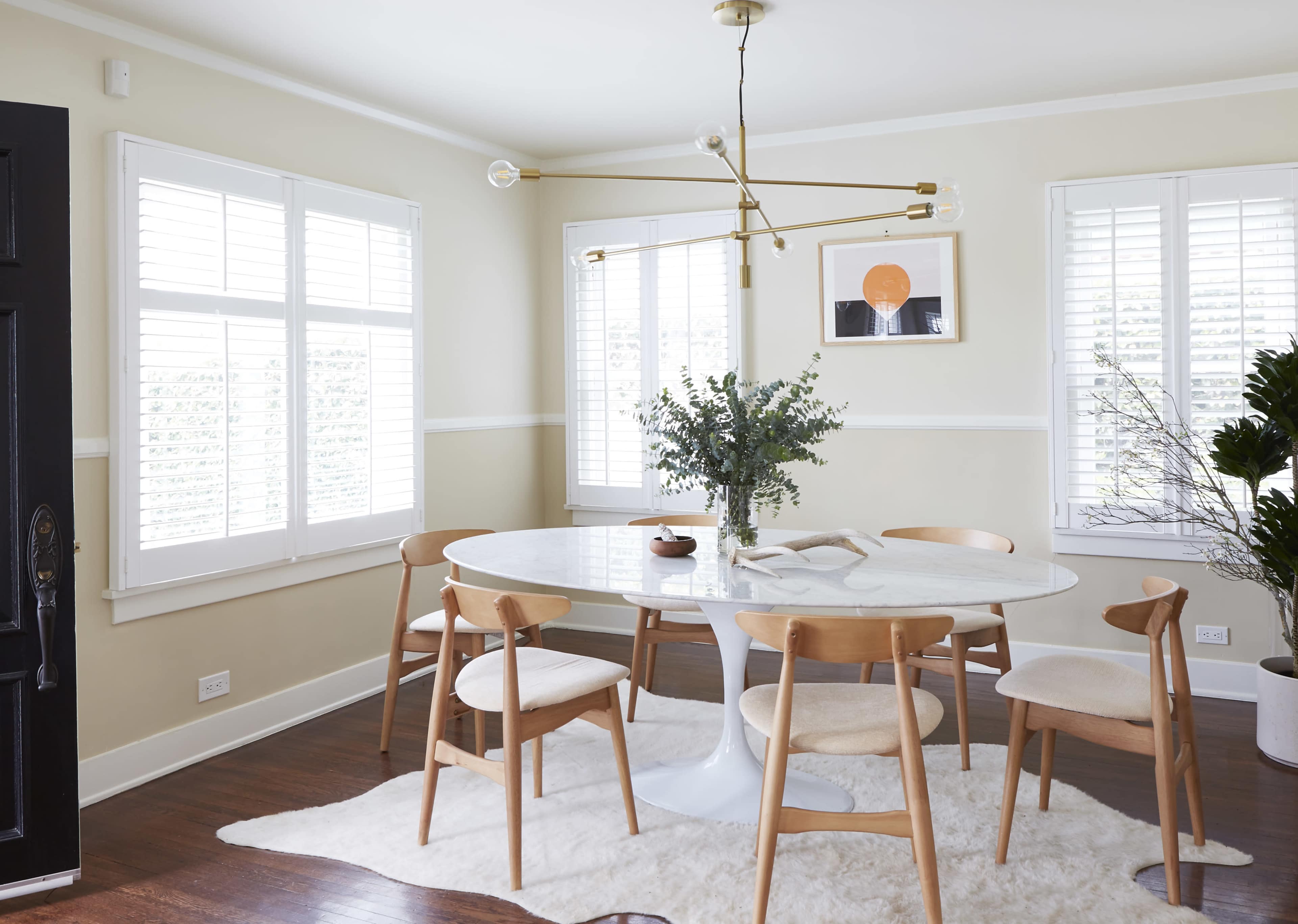 A round white dining table with wooden chairs is set in a well-lit room featuring large windows and minimal decor.