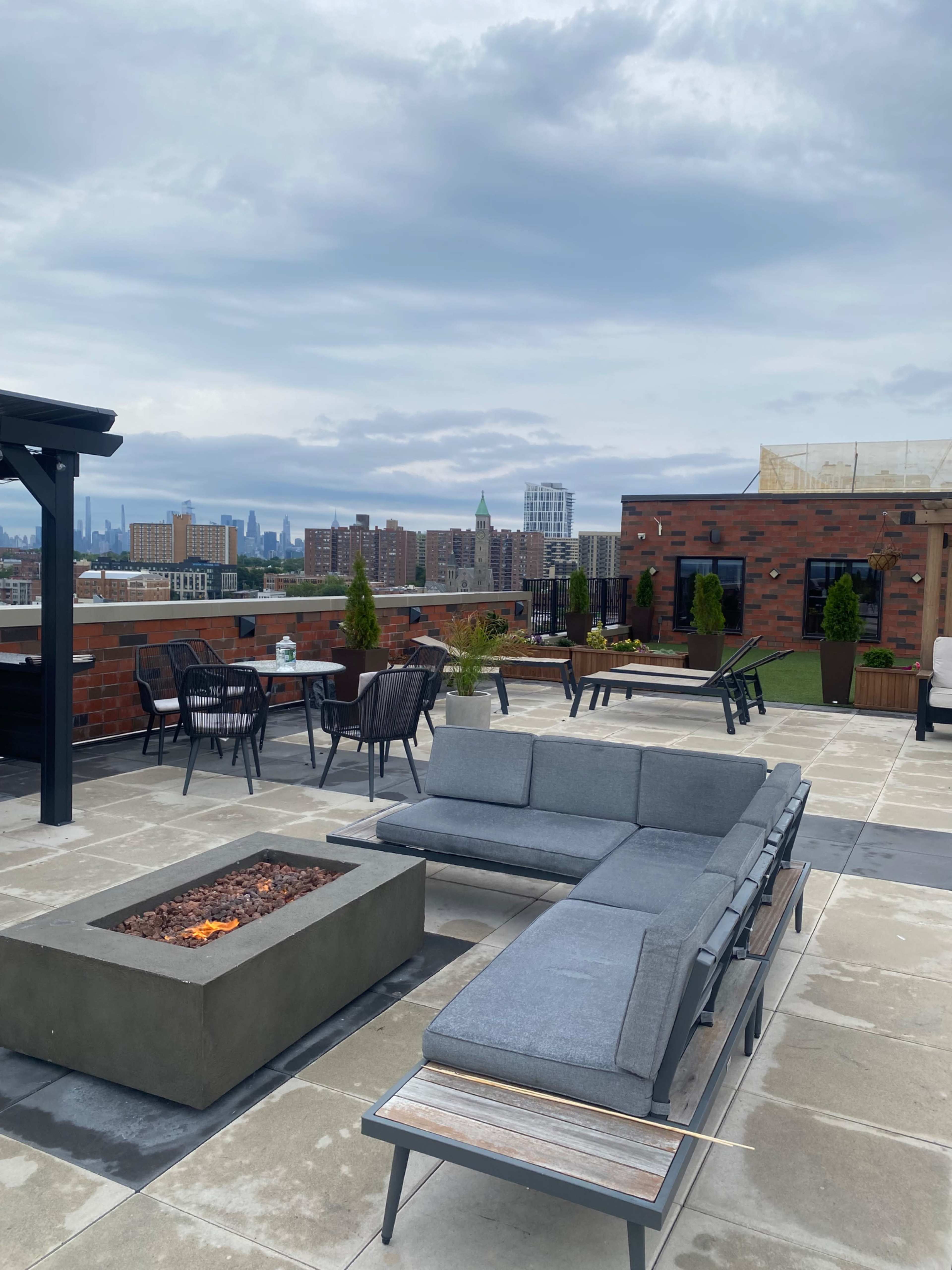 The image shows a rooftop terrace featuring modern seating, a fire pit, and a view of a city skyline with overcast skies.