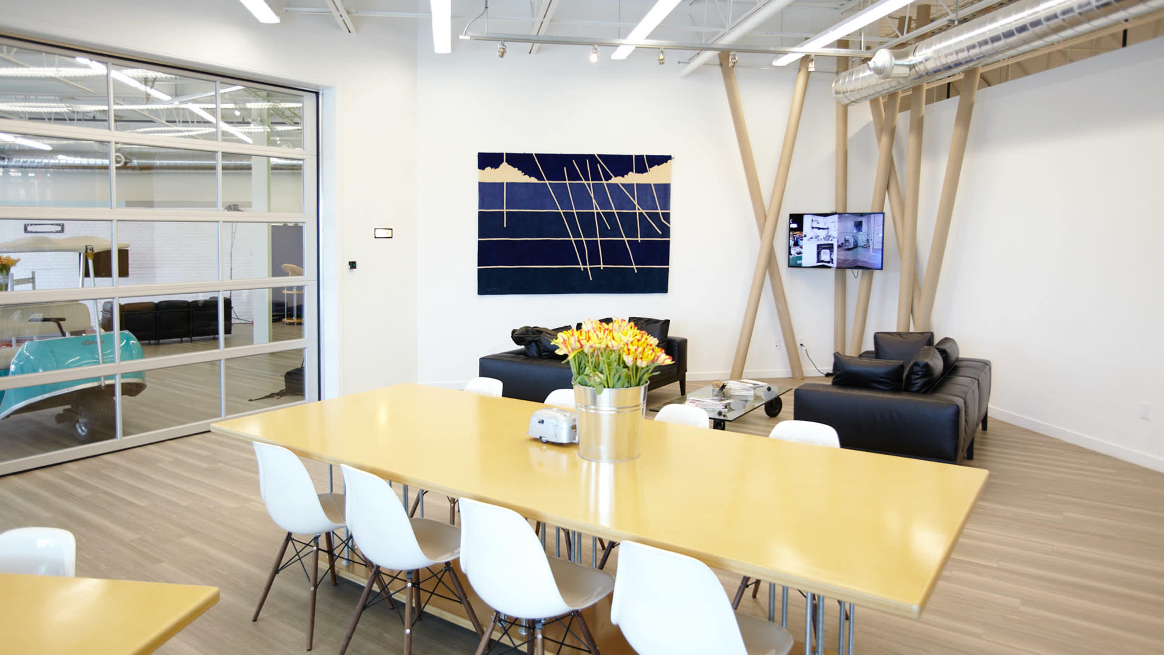 A modern, open office space features a long yellow table with white chairs, a black sofa area, and a large wall art piece.