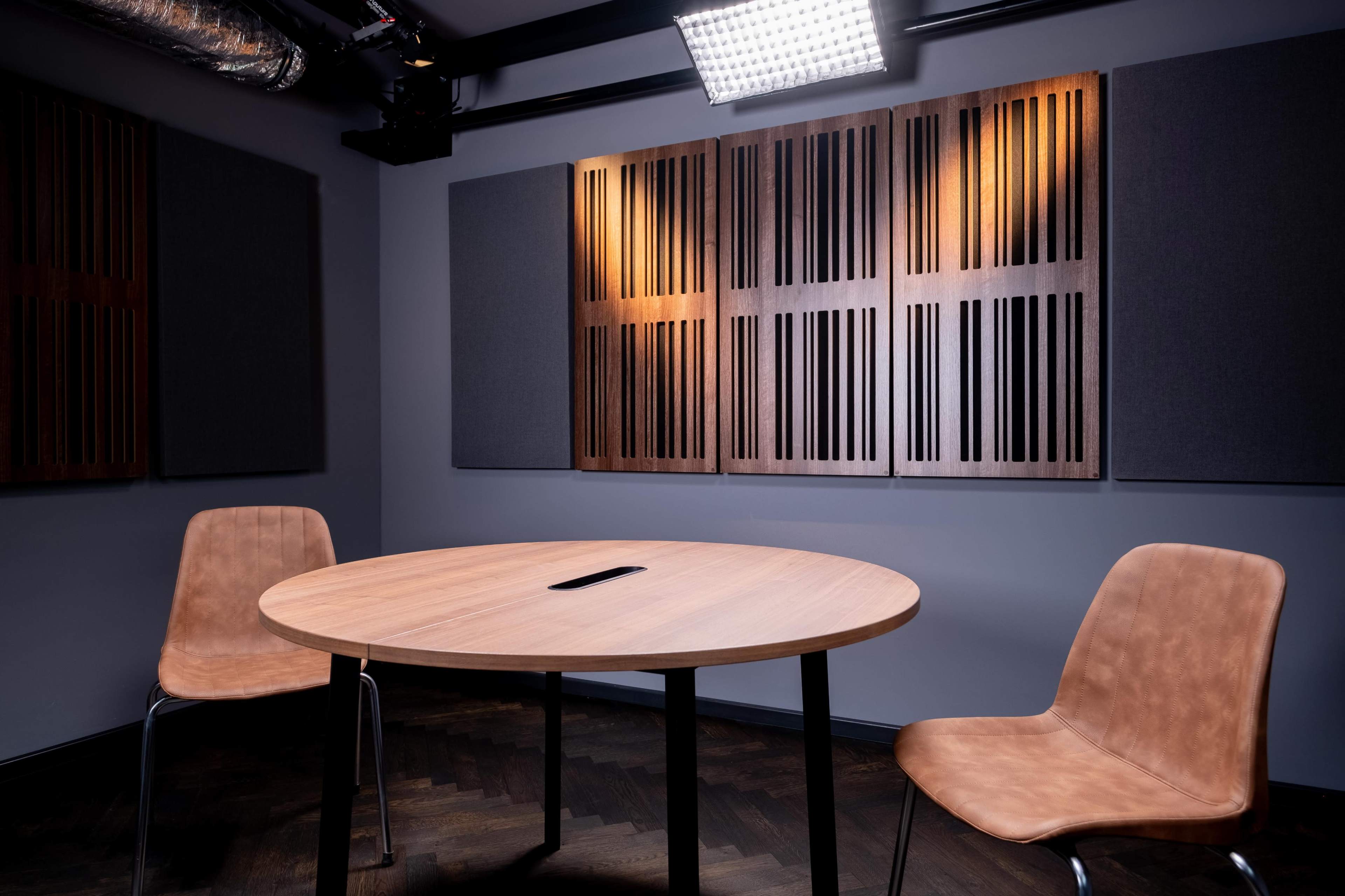 A circular wooden table is set in a soundproof room, accompanied by two tan chairs and sound-absorbing panels on the walls.