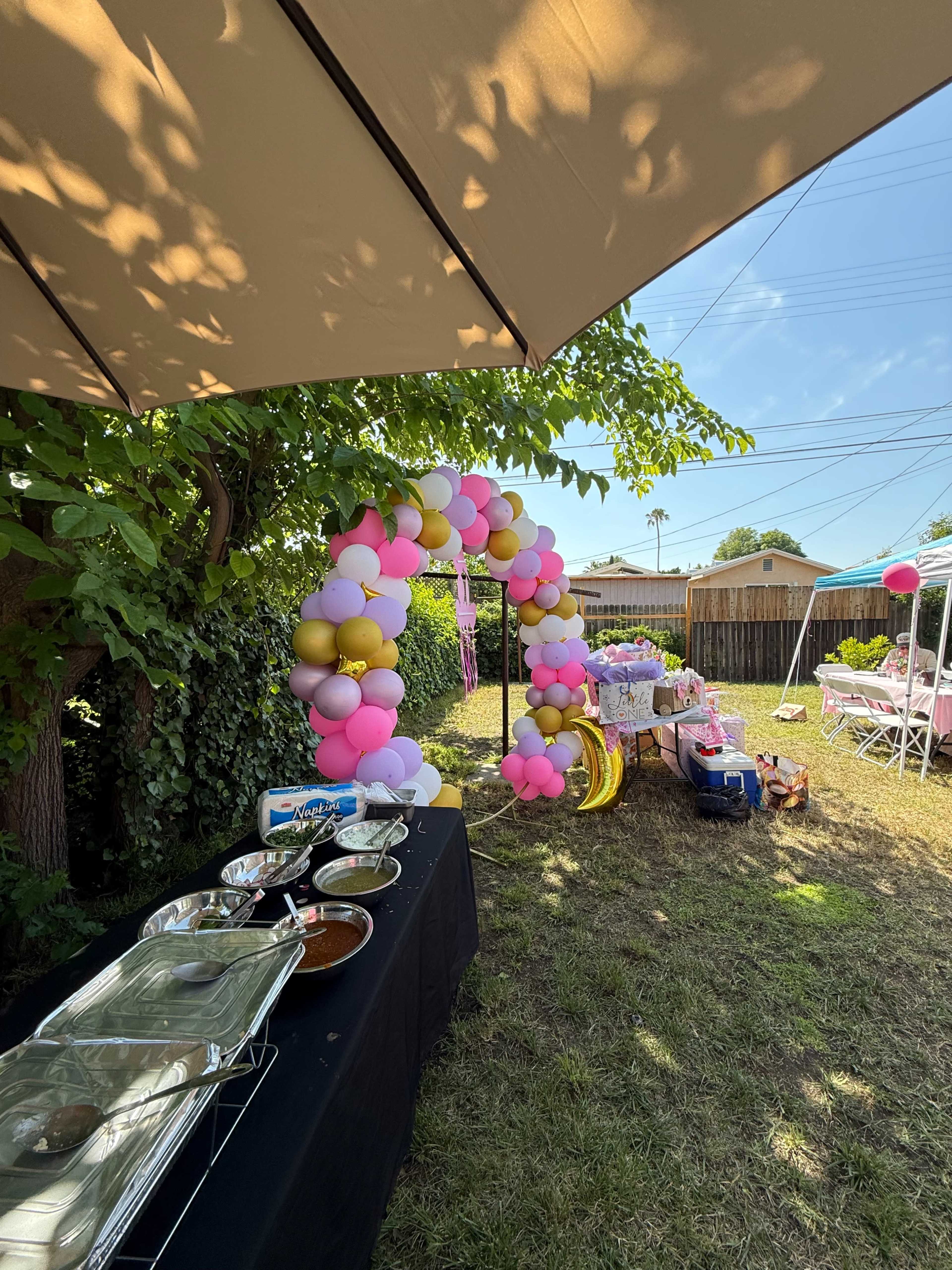 A colorful balloon arch is set up in a backyard next to a table with buffet-style food and various decorations for a celebration.