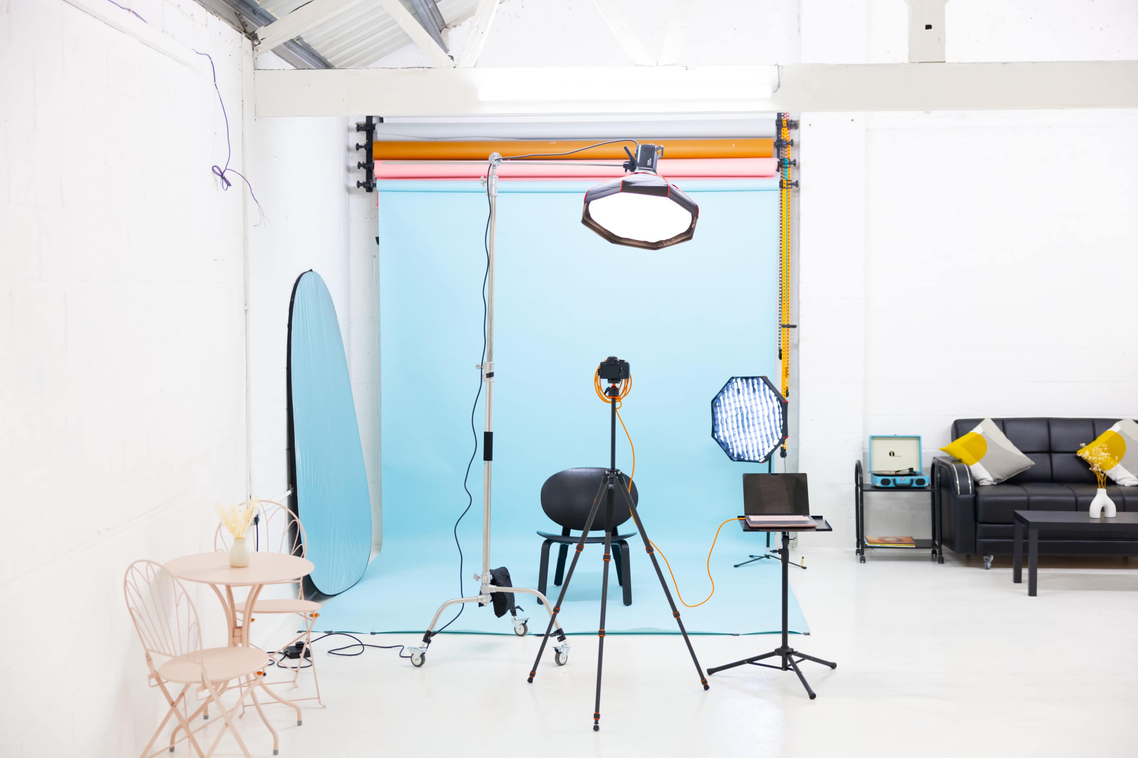 A photography studio features lighting equipment, a blue backdrop, a black chair, and a small table with chairs.