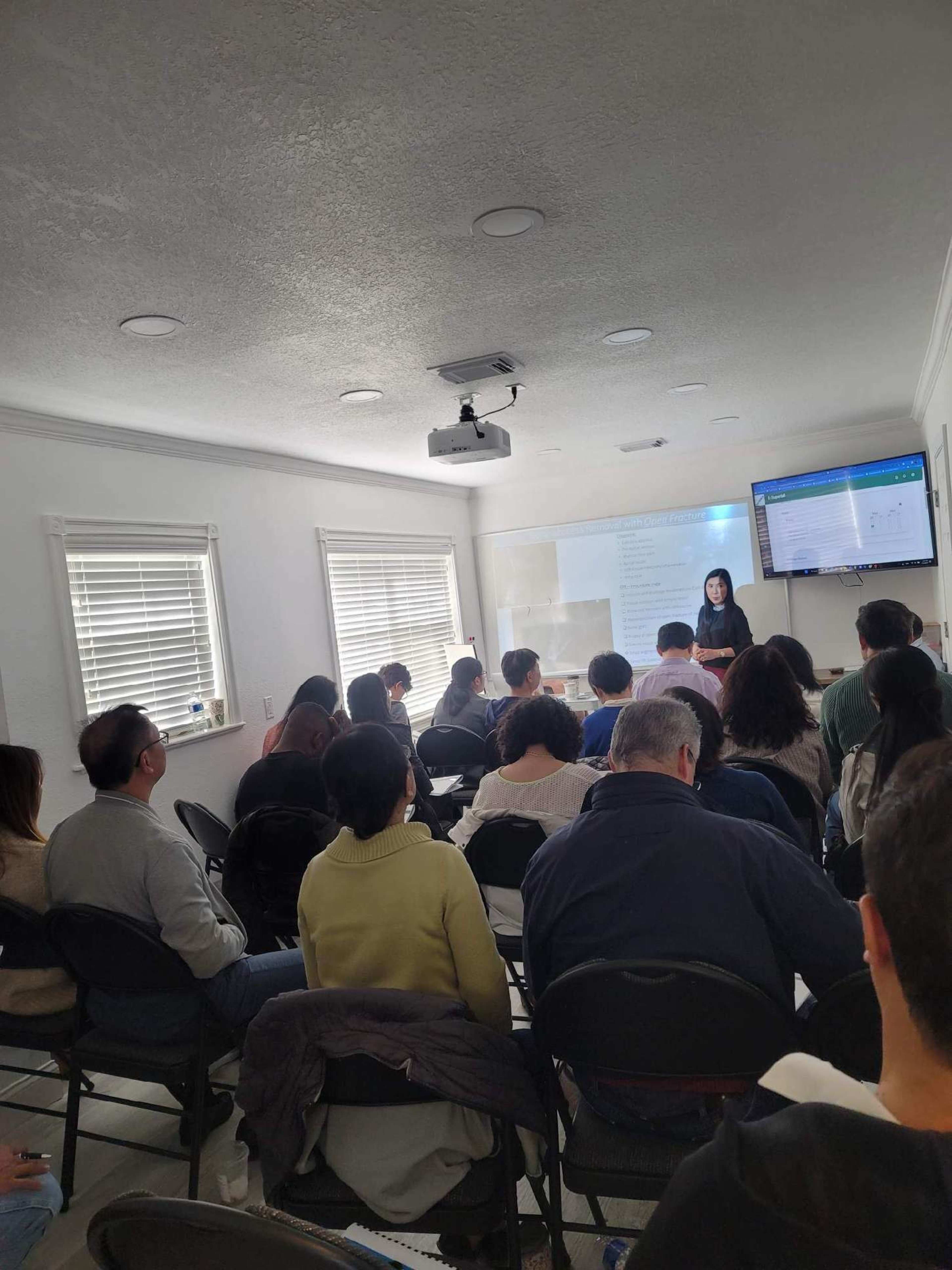 A group of people is seated in chairs, facing a woman who is presenting in front of a screen in a well-lit room.