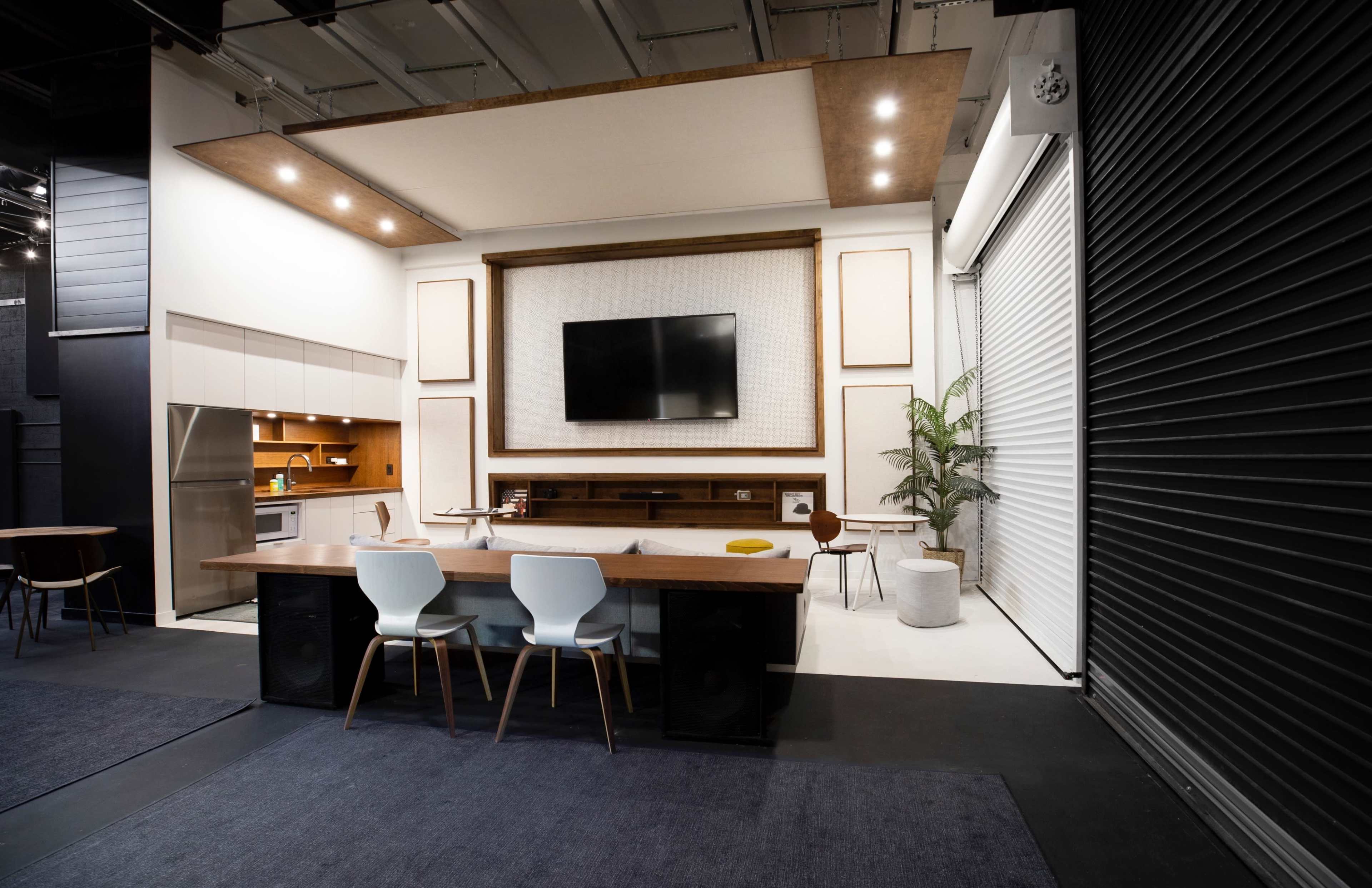 The image shows a modern indoor space featuring a kitchen area, a large television mounted on a wall, a dining table with chairs, and a potted plant.