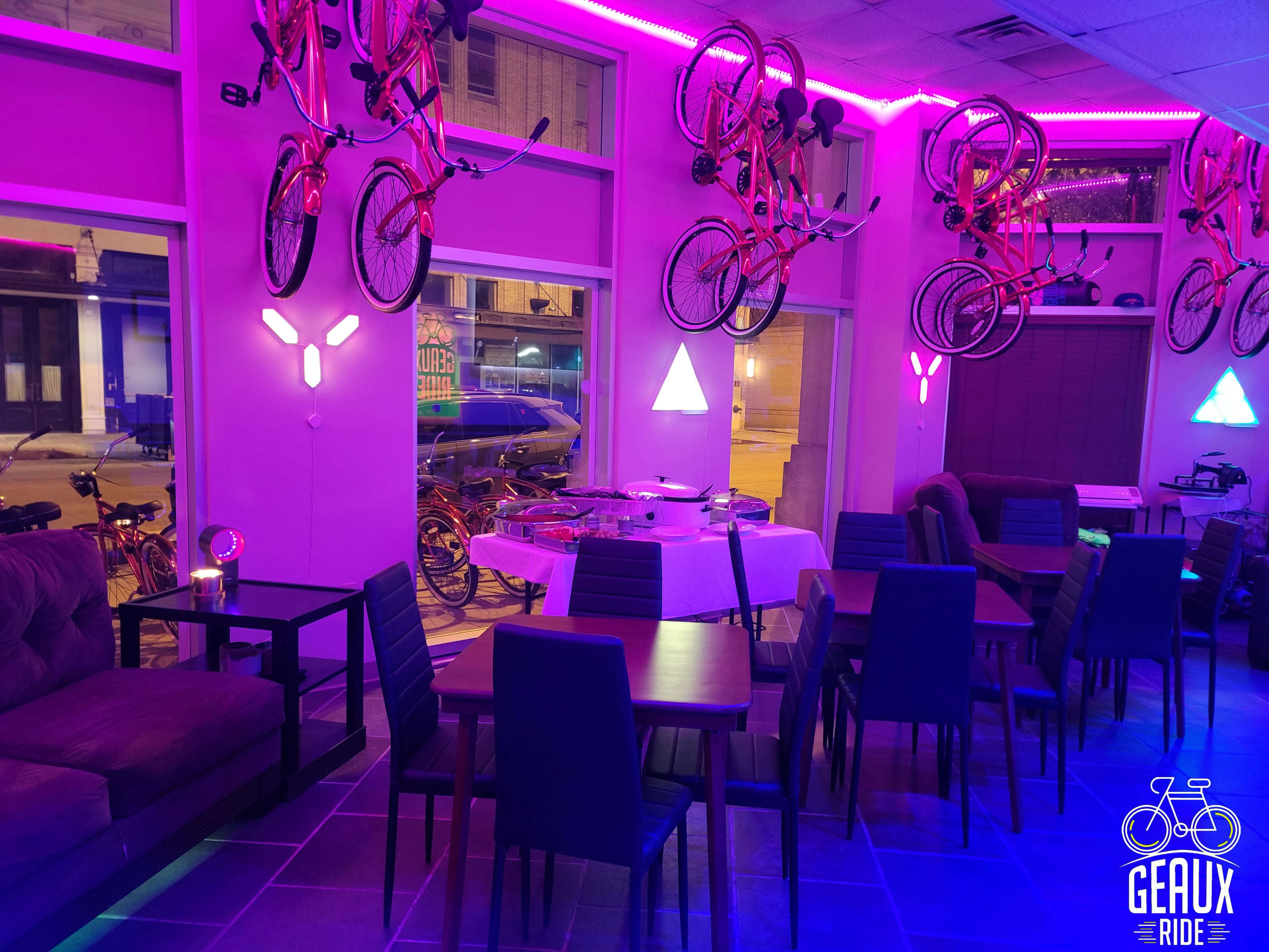 The interior of a cafe features purple lighting, tables, and chairs, with bicycles hanging on the walls above.