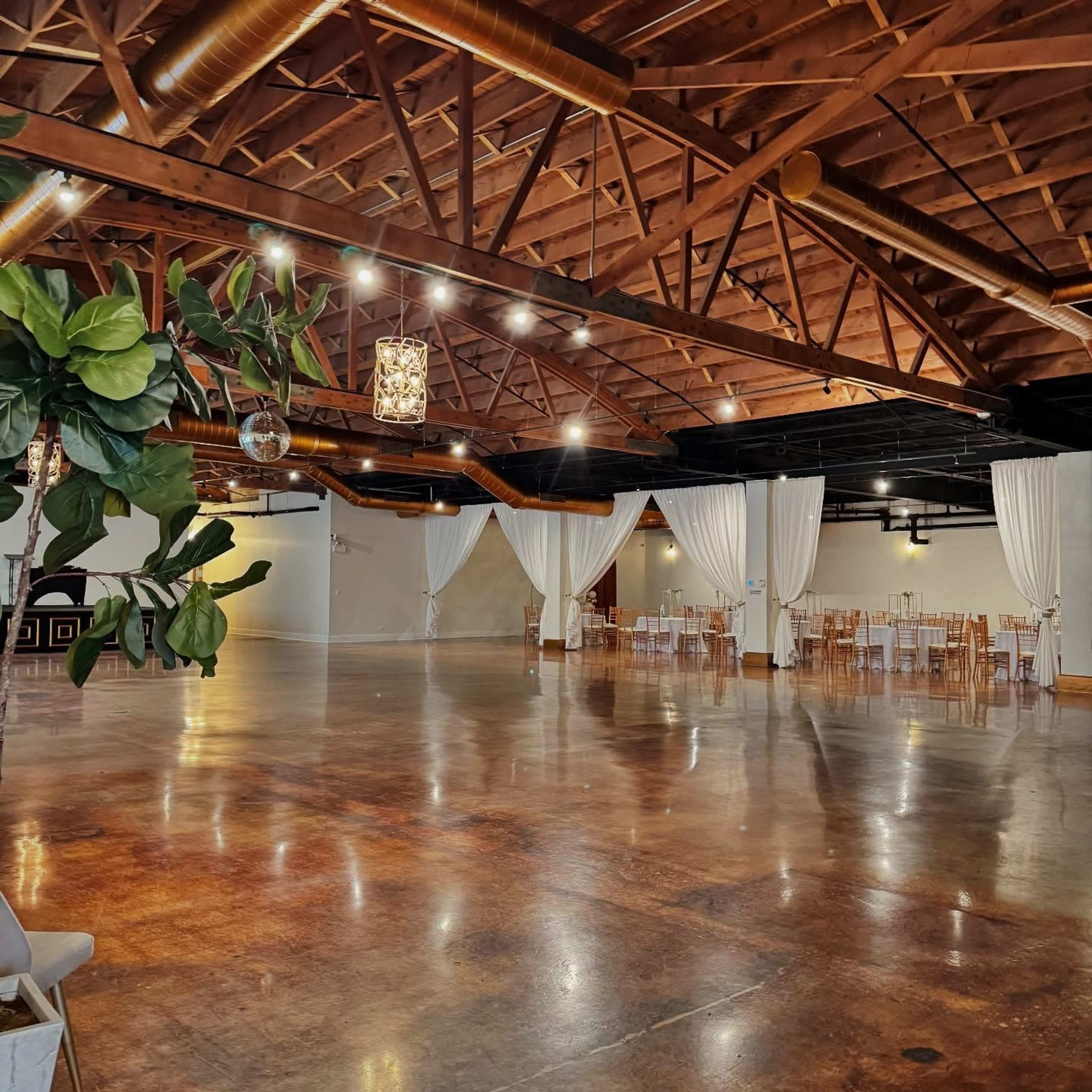 The image shows a spacious event venue with exposed wooden beams, polished floors, and white drapes separating seating areas.