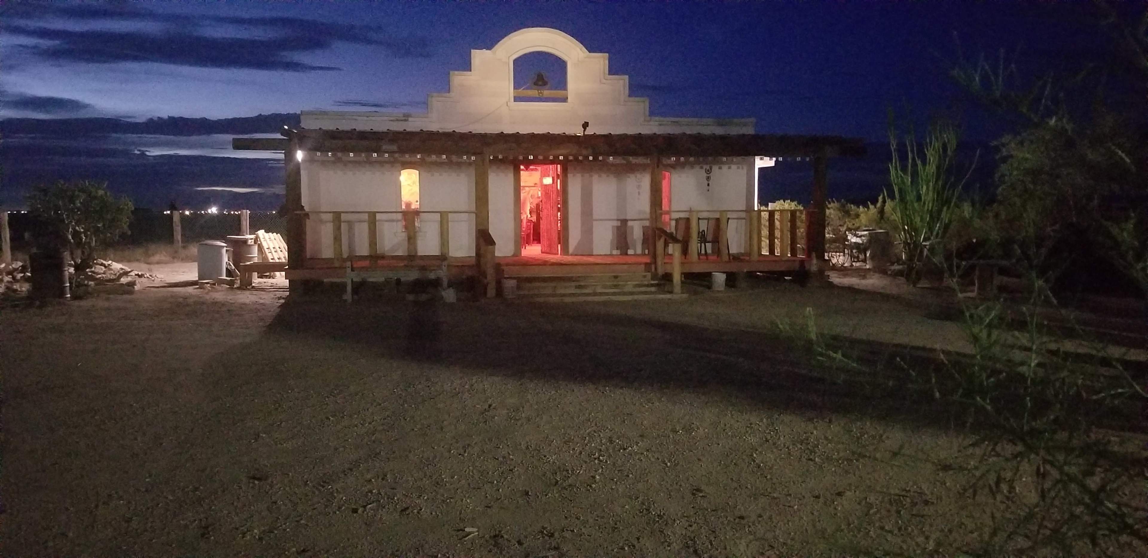 A brightly lit building with a porch stands alone in a desert landscape during twilight.