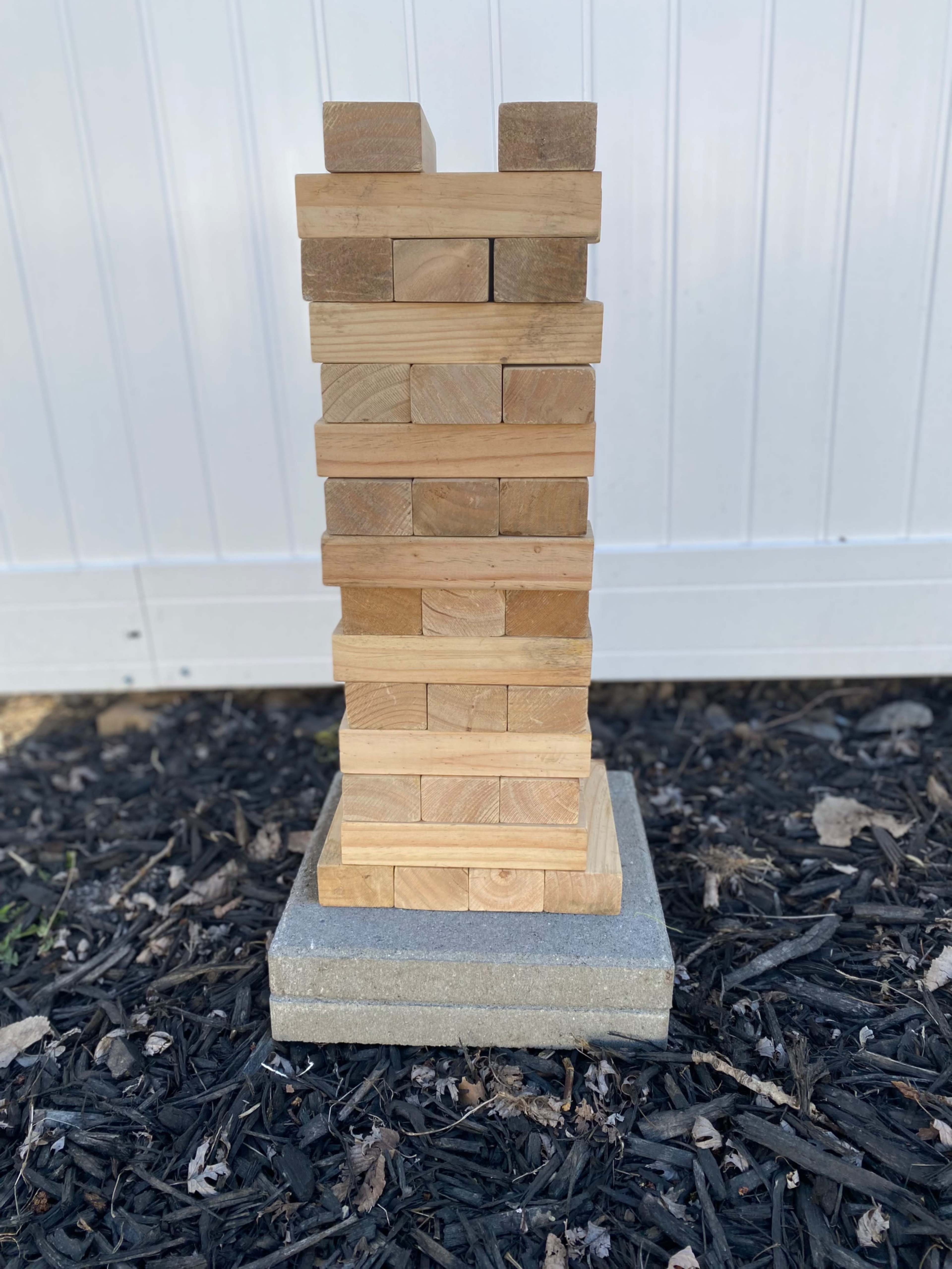 A tower of wooden blocks is stacked neatly on a concrete base, resting on a bed of dark mulch.