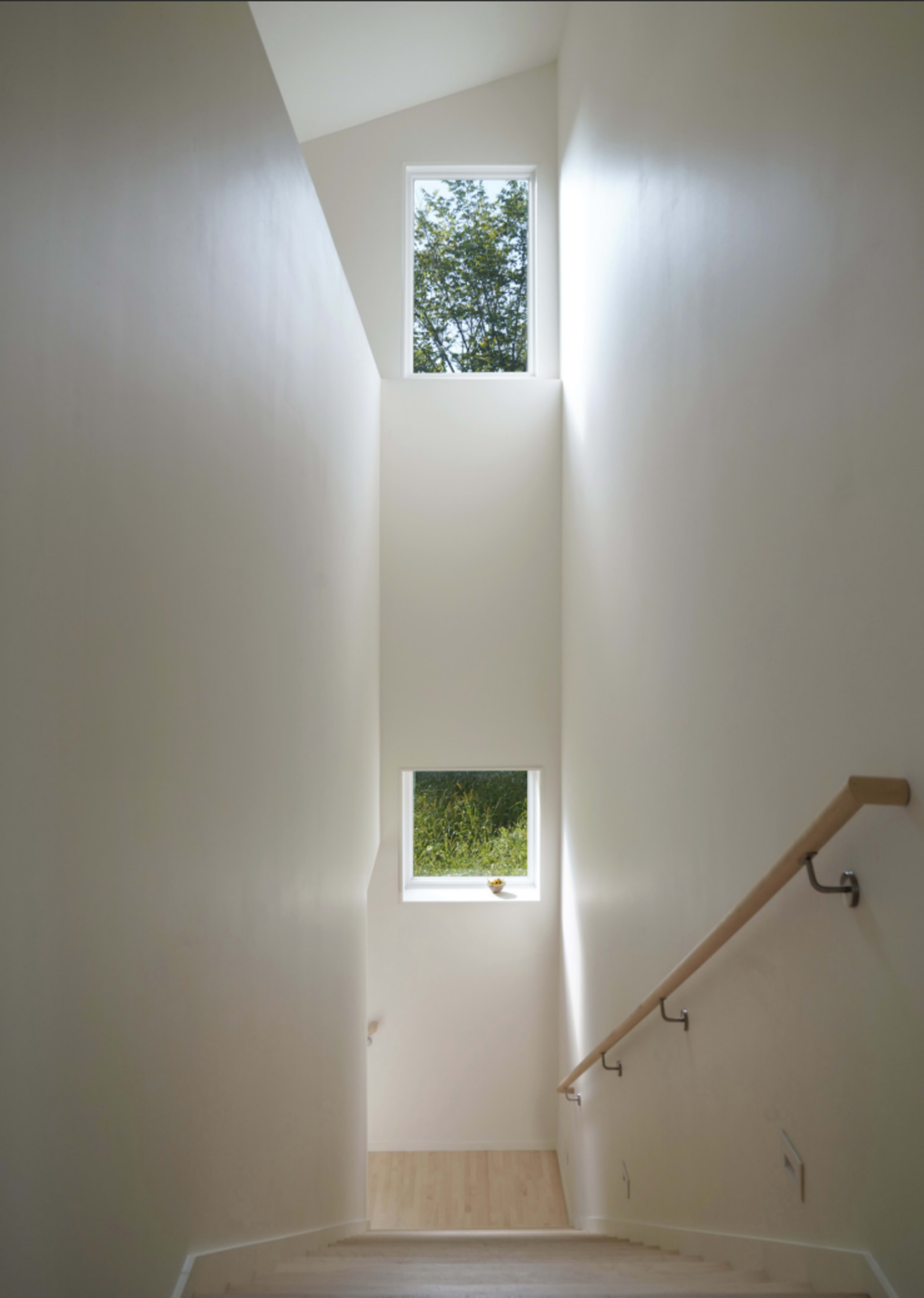 The image shows a light-filled staircase with two square windows at the top and bottom, framed by white walls and a wooden railing.