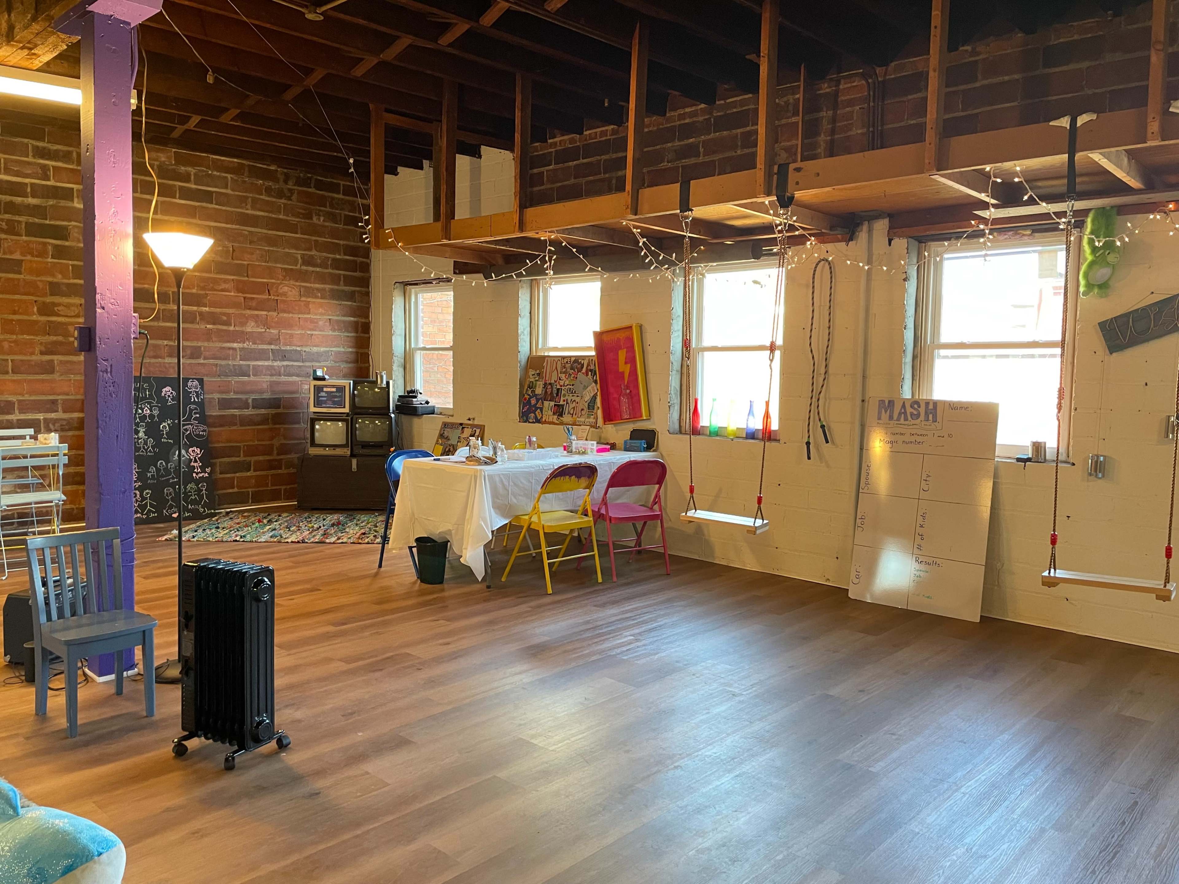 A spacious room features a table with colorful chairs, a lamp, and wall decorations, all under exposed wooden beams.